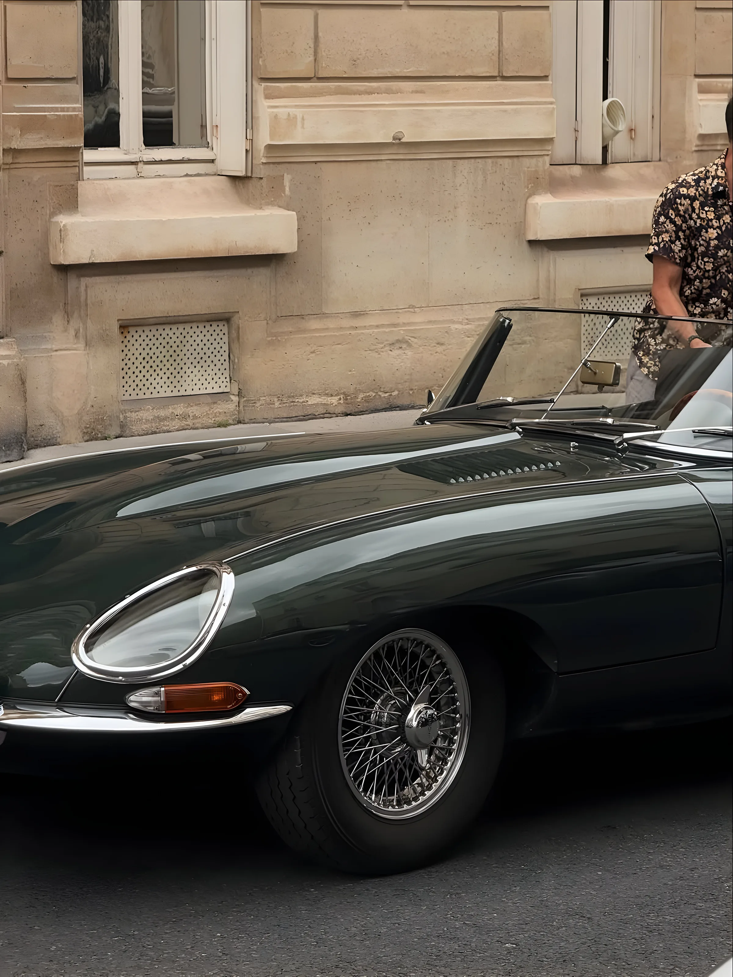 Dark green vintage convertible sports car with wire wheels parked by a stone facade, a man in a floral shirt beside it.