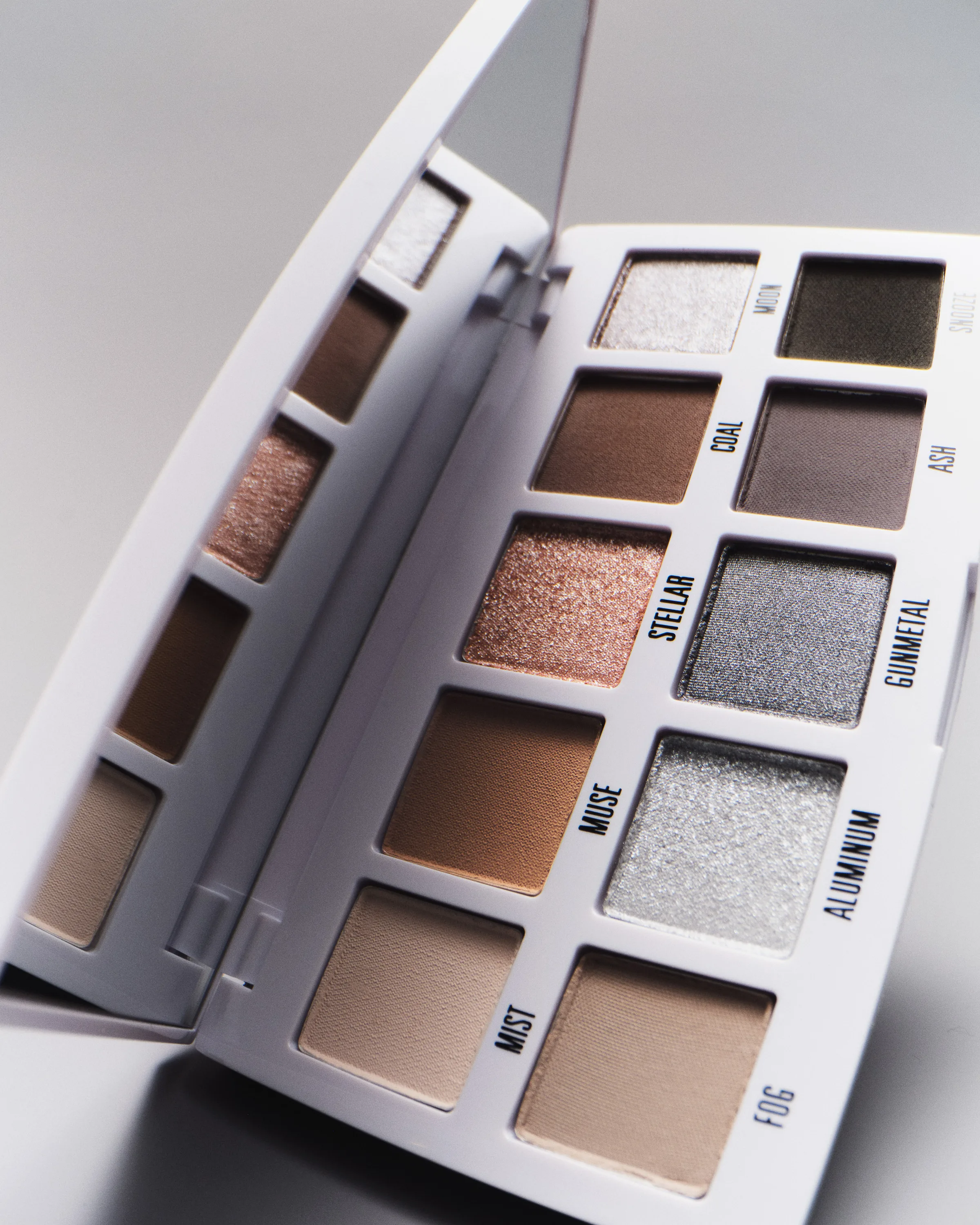 Open white eyeshadow palette with smoky neutral and metallic shades, mirror lid raised, on grey, for Kylie Cosmetics.