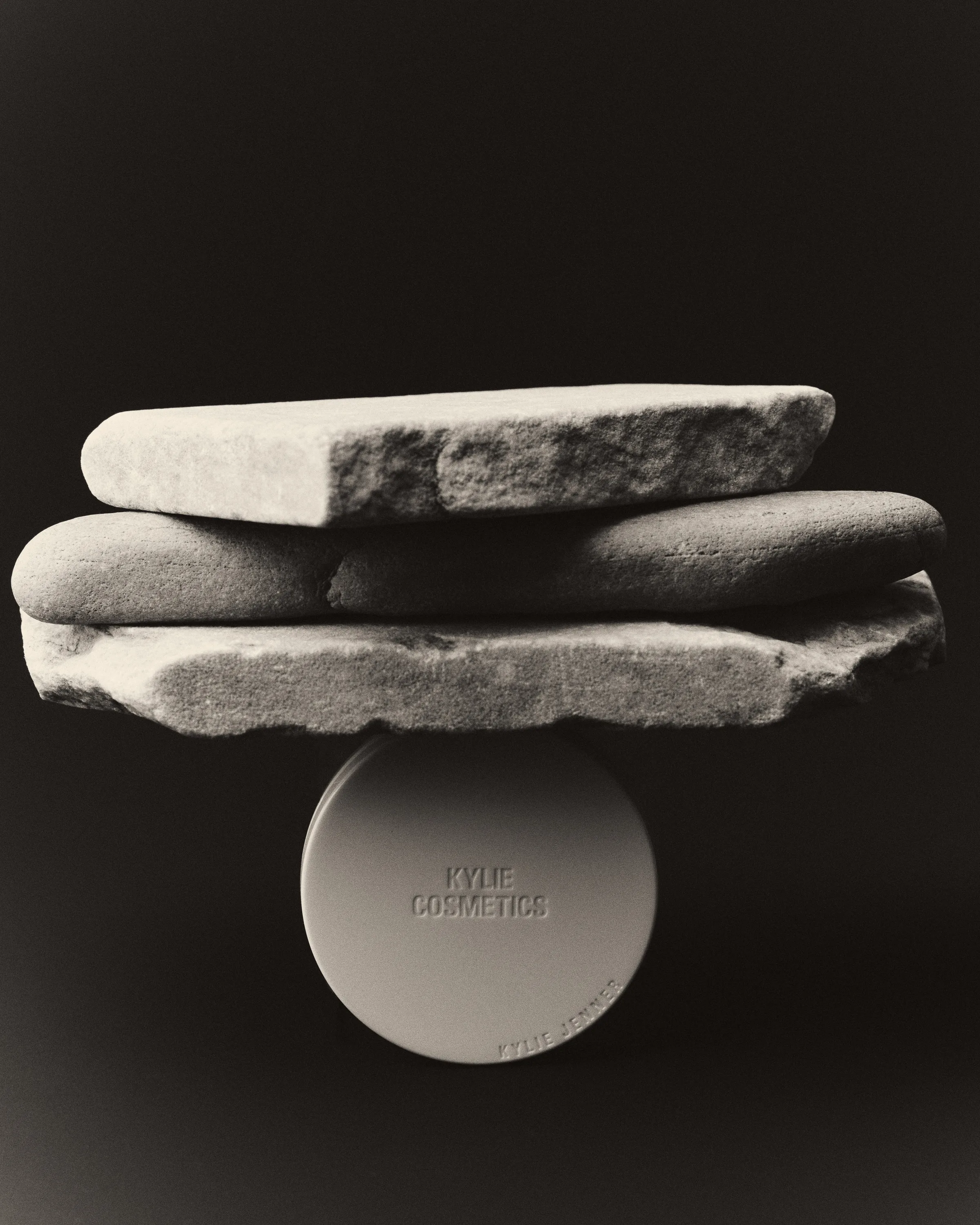 Black and white still life of a round compact balancing under three stacked flat stones, for Kylie Cosmetics.