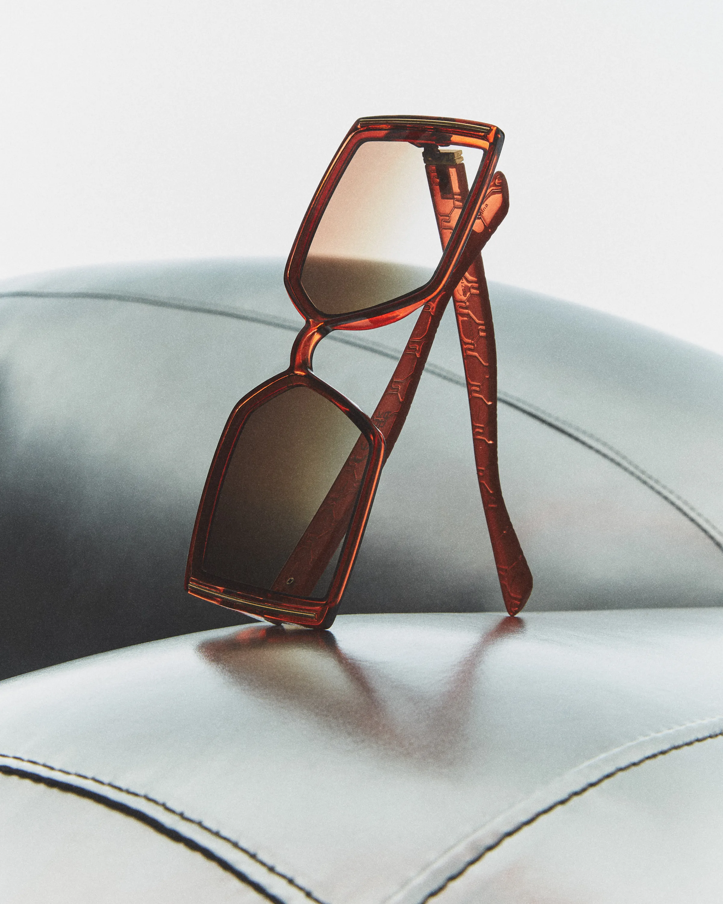 Translucent red rectangular sunglasses with embossed arms propped on a grey leather chair, for Sam Edelman.