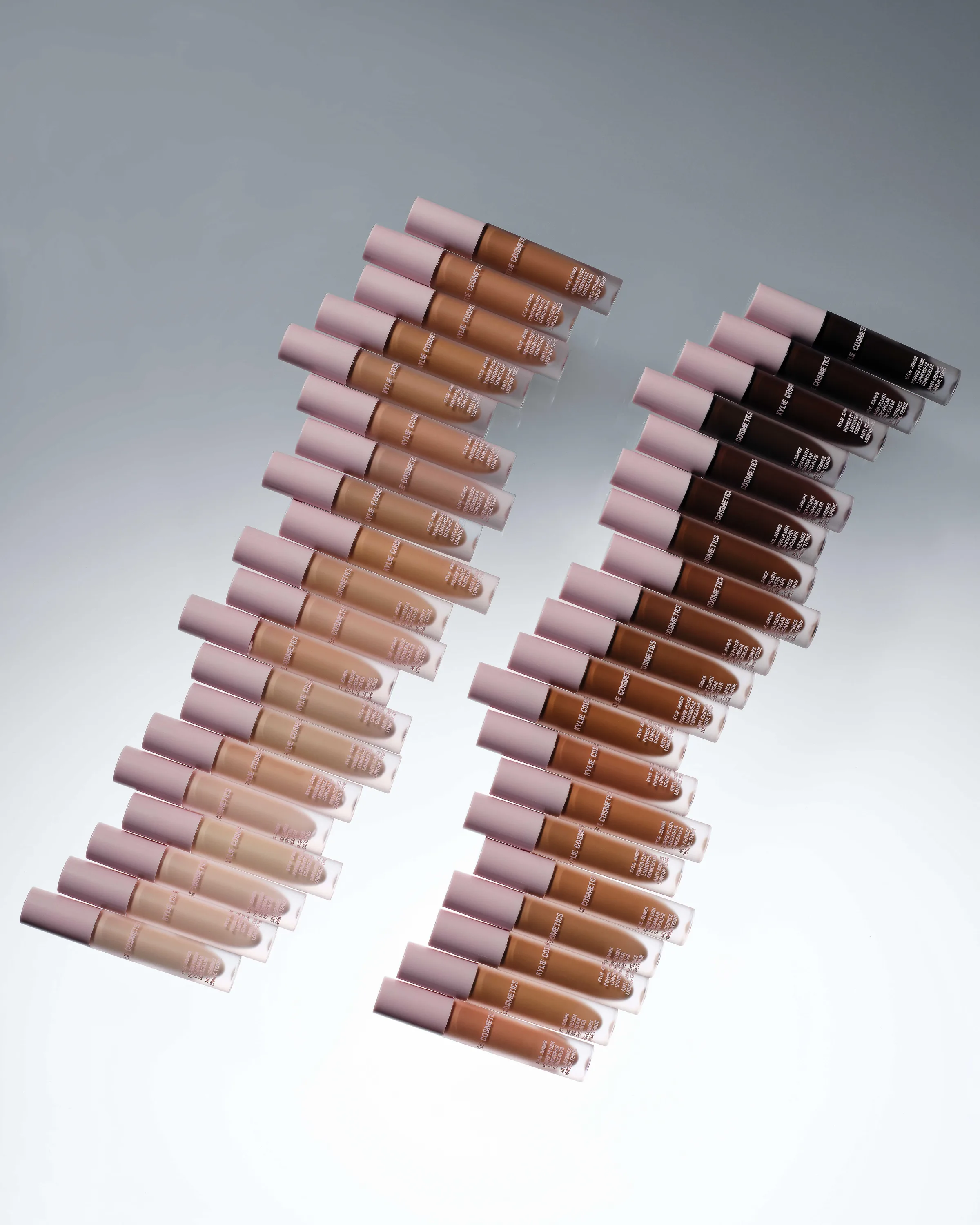 Two diagonal rows of concealer tubes with pink caps, arranged light to deep shades on a grey gradient, for Kylie Cosmetics.