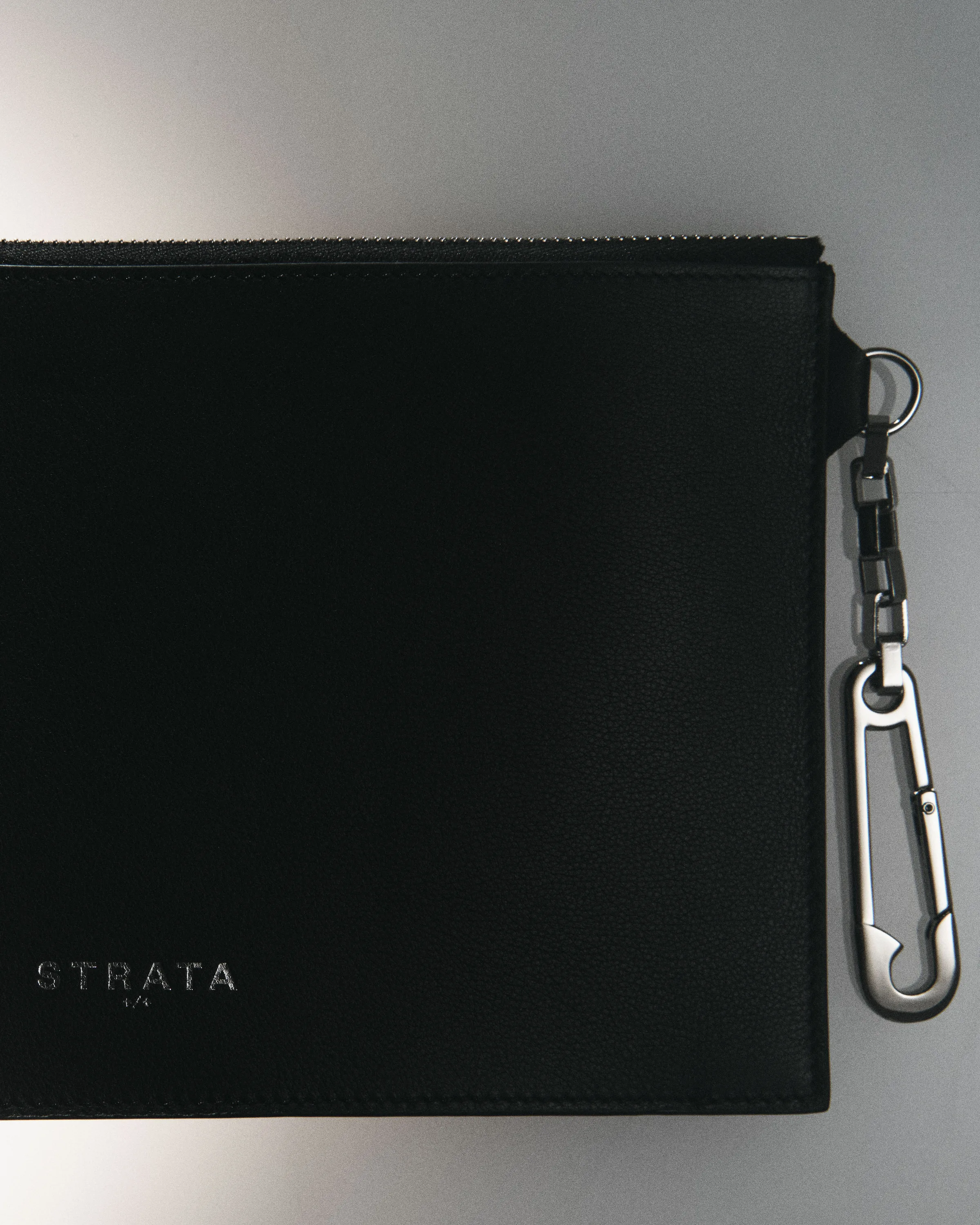Black leather zip clutch with silver logo and safety-pin clip keychain on a grey gradient backdrop, for Strata.