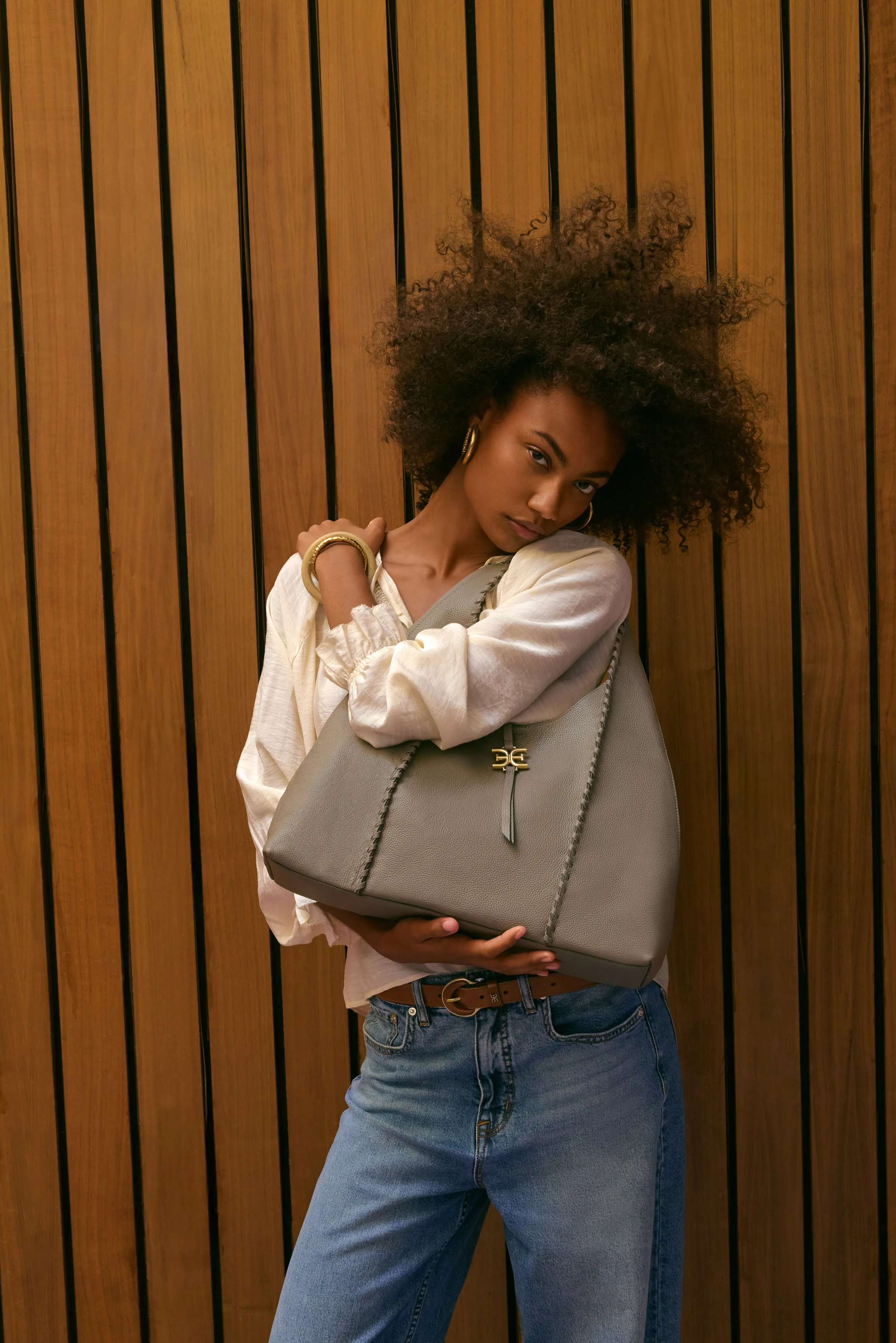 Model hugging a grey-green leather tote with gold hardware, cream blouse and jeans, against wood slat wall, for Sam Edelman.