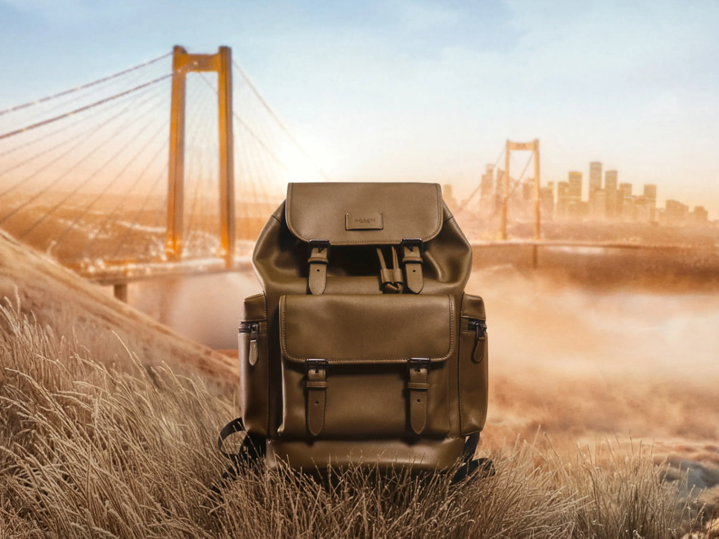 AI image of an olive leather backpack on golden grass before a misty suspension bridge and skyline, for Coach.