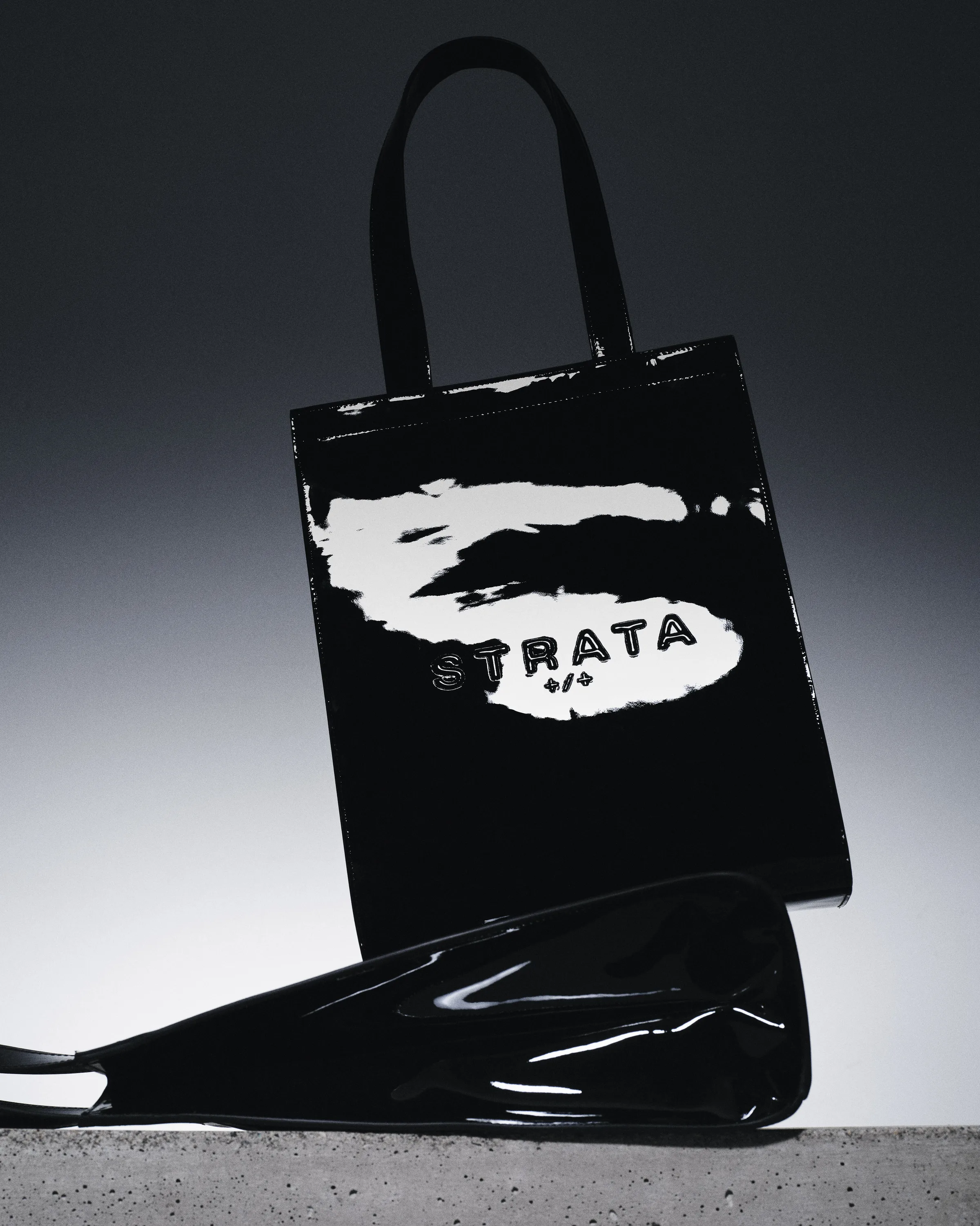 Black patent leather Carmen tote with embossed logo, a second bag lying on concrete against a dark gradient, for Strata.