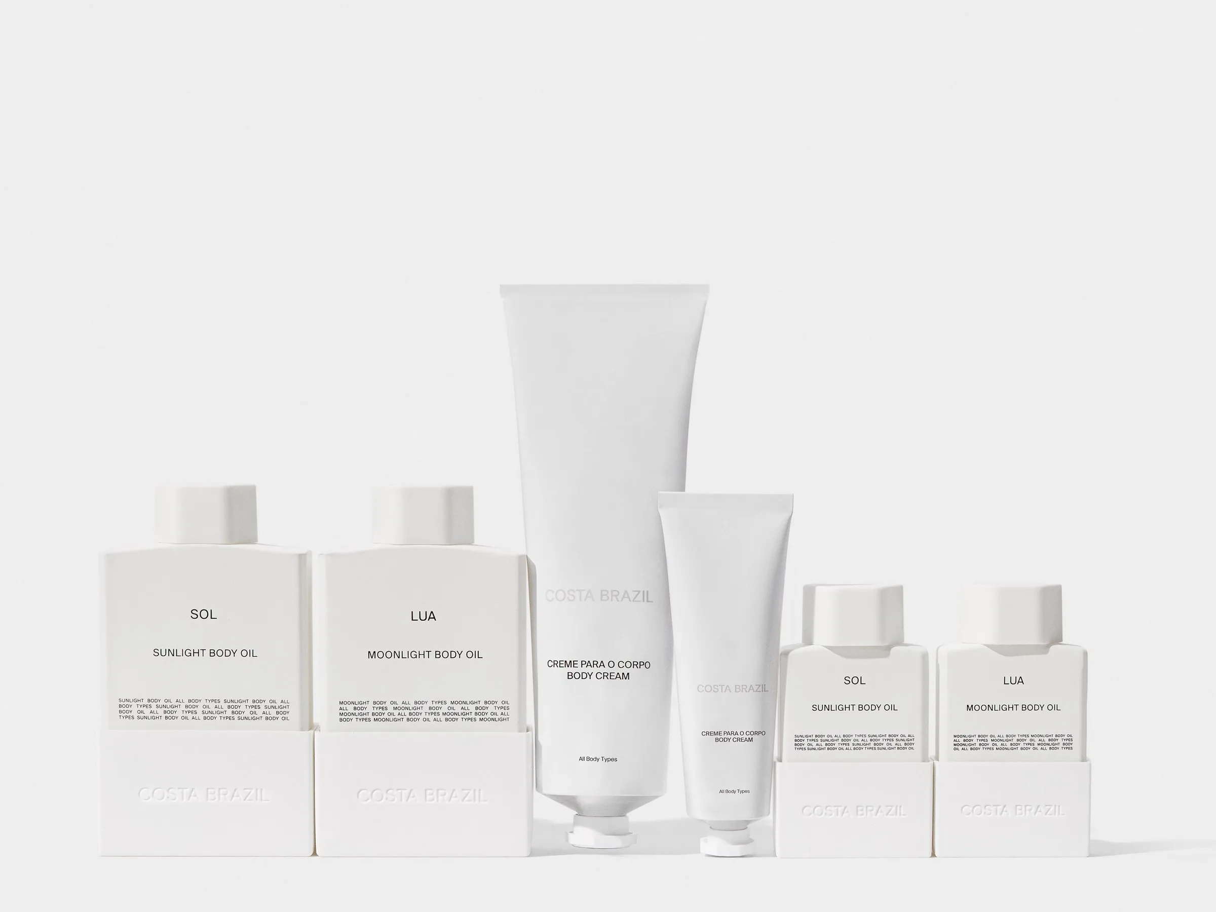 Six white bottles and tubes of Sol and Lua body oils and body cream lined up on a light grey background, for Costa Brazil.