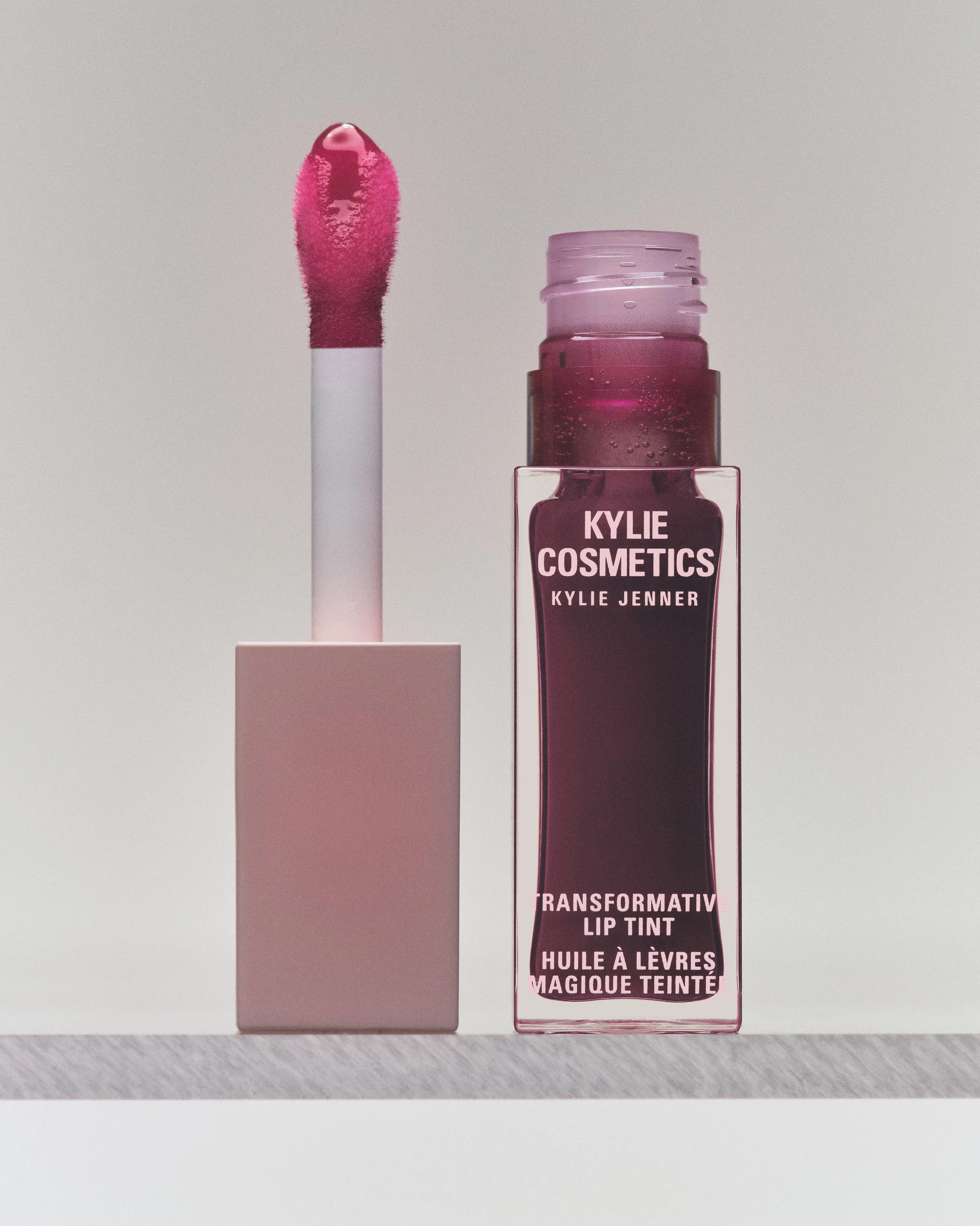 Plum transformative lip tint bottle beside its pink doe-foot applicator on a grey ledge, for Kylie Cosmetics.