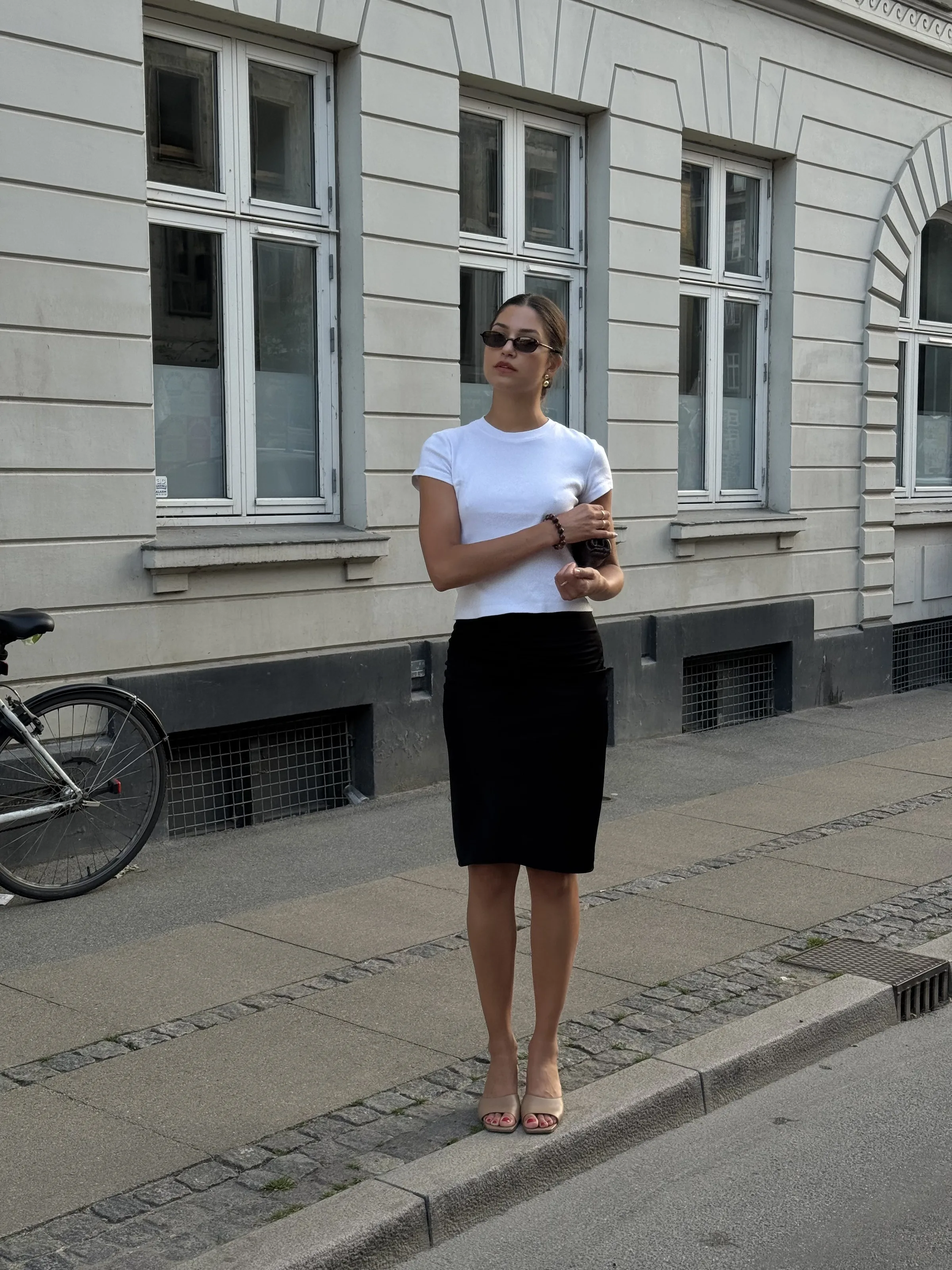 Woman in sunglasses, white tee and black pencil skirt standing on a cobblestone sidewalk outside a grey building.