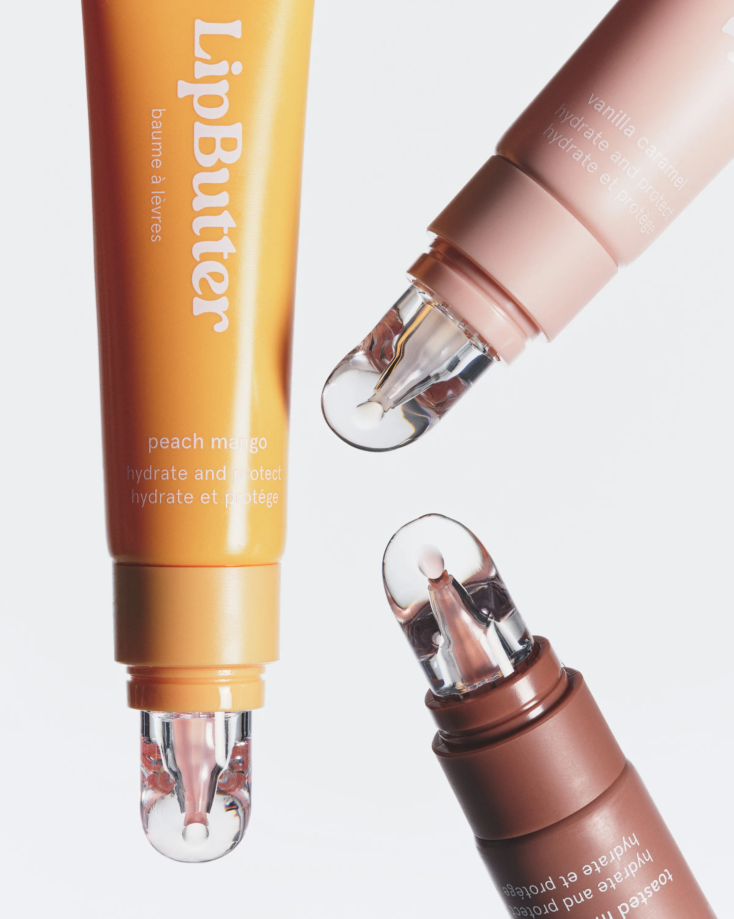 Three Lip Butter tubes in peach mango, vanilla caramel and toasted shades with clear tips on white, for Kylie Cosmetics.