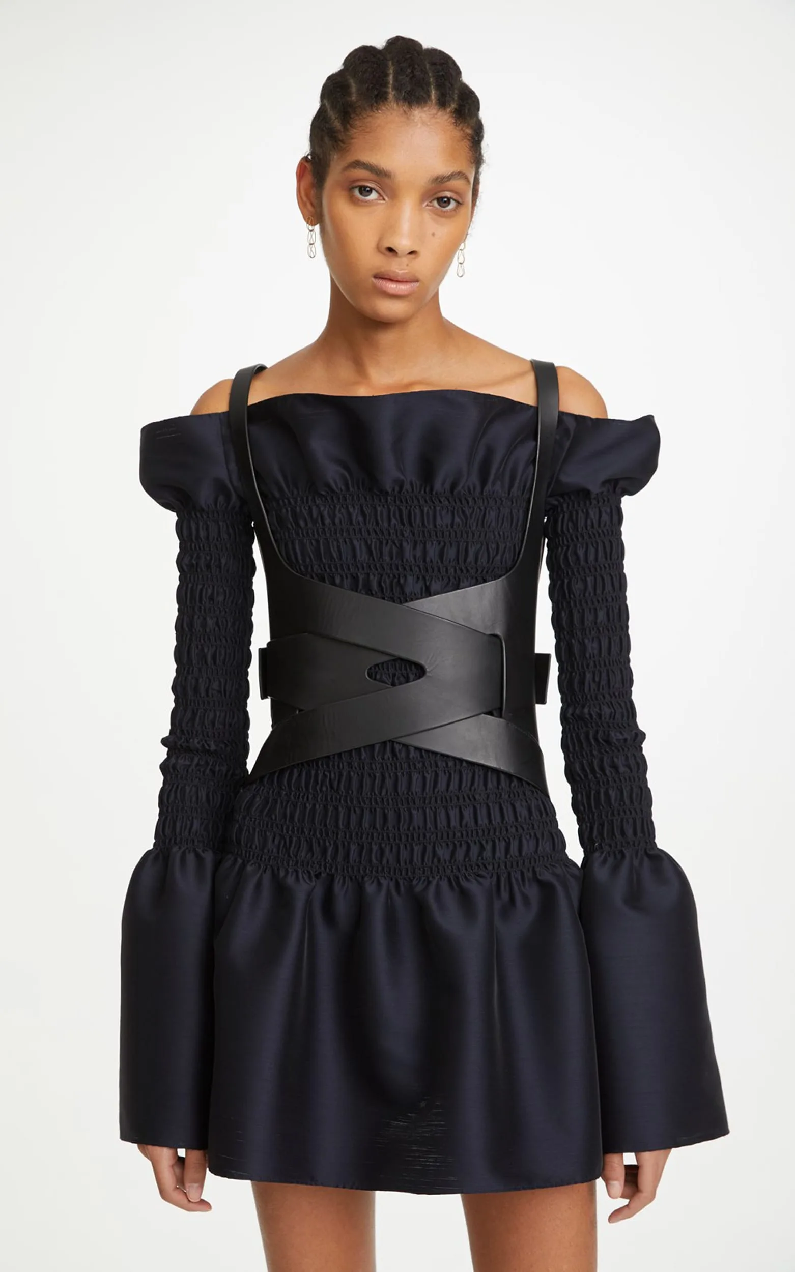 Model in a navy smocked off-shoulder mini dress with bell sleeves and black leather harness on white, for Dion Lee.