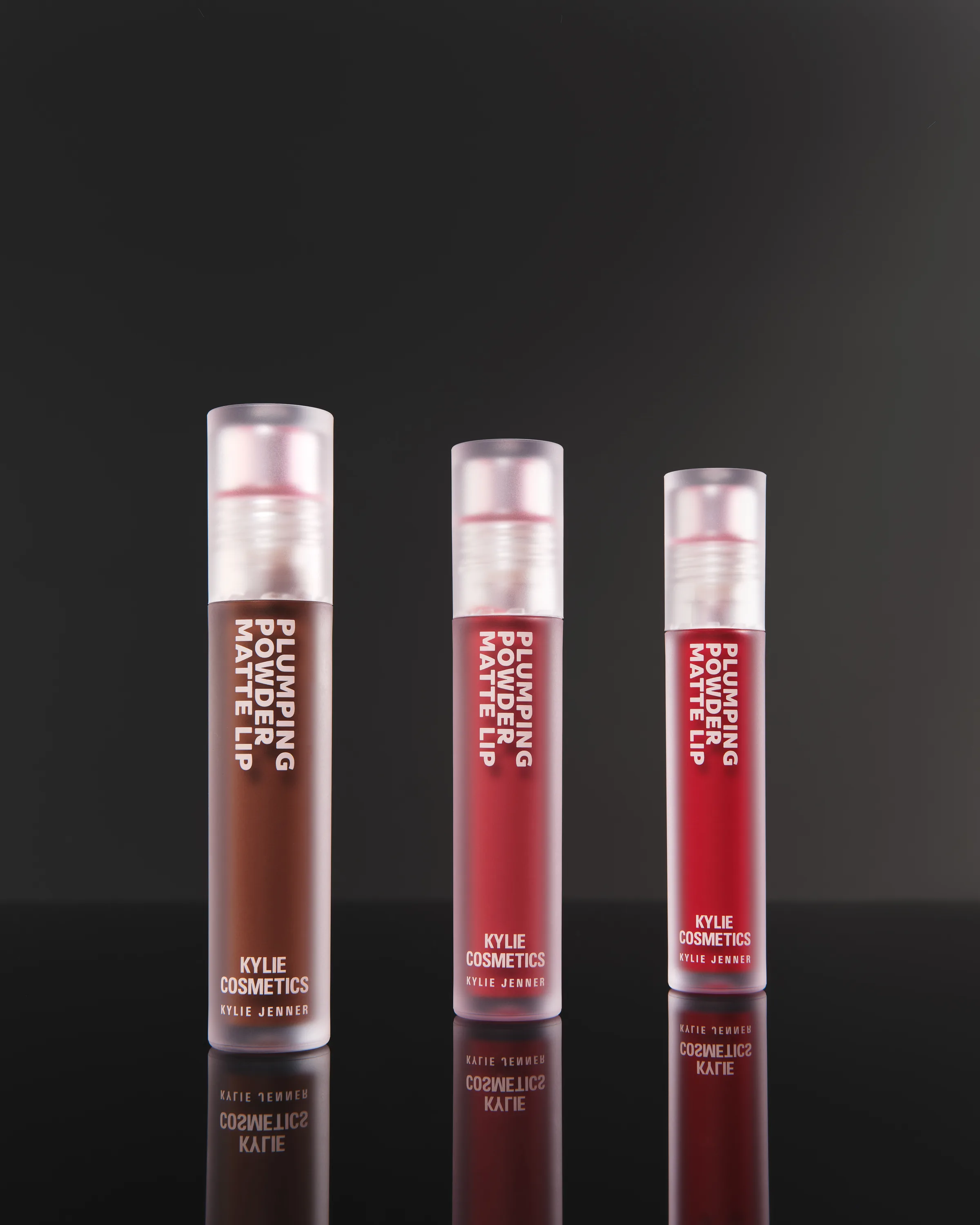 Three Plumping Powder Matte Lip tubes in brown, red and crimson reflected on a glossy black surface, for Kylie Cosmetics.