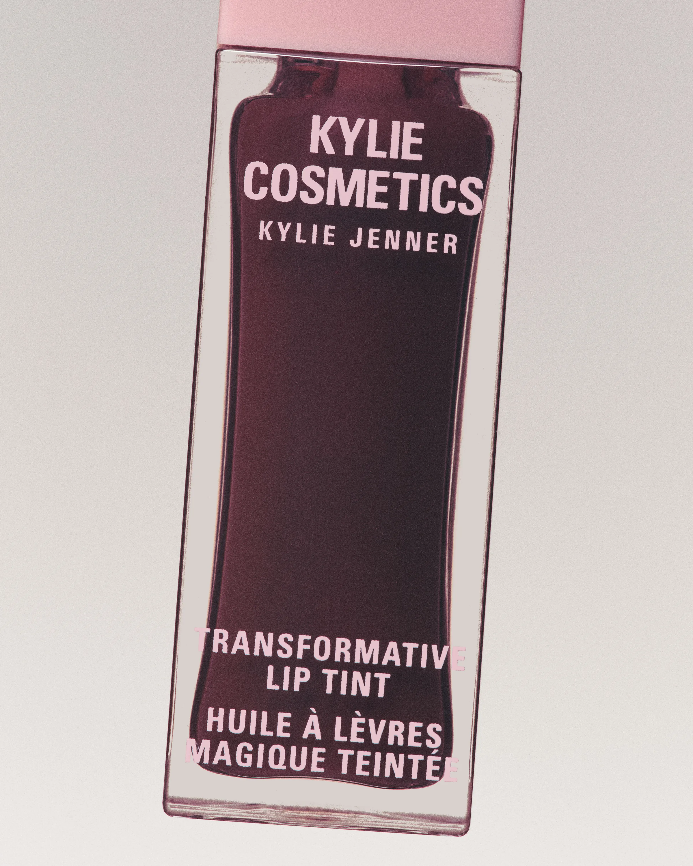 Plum Transformative Lip Tint bottle with pink cap, tilted against a soft grey backdrop, for Kylie Cosmetics.