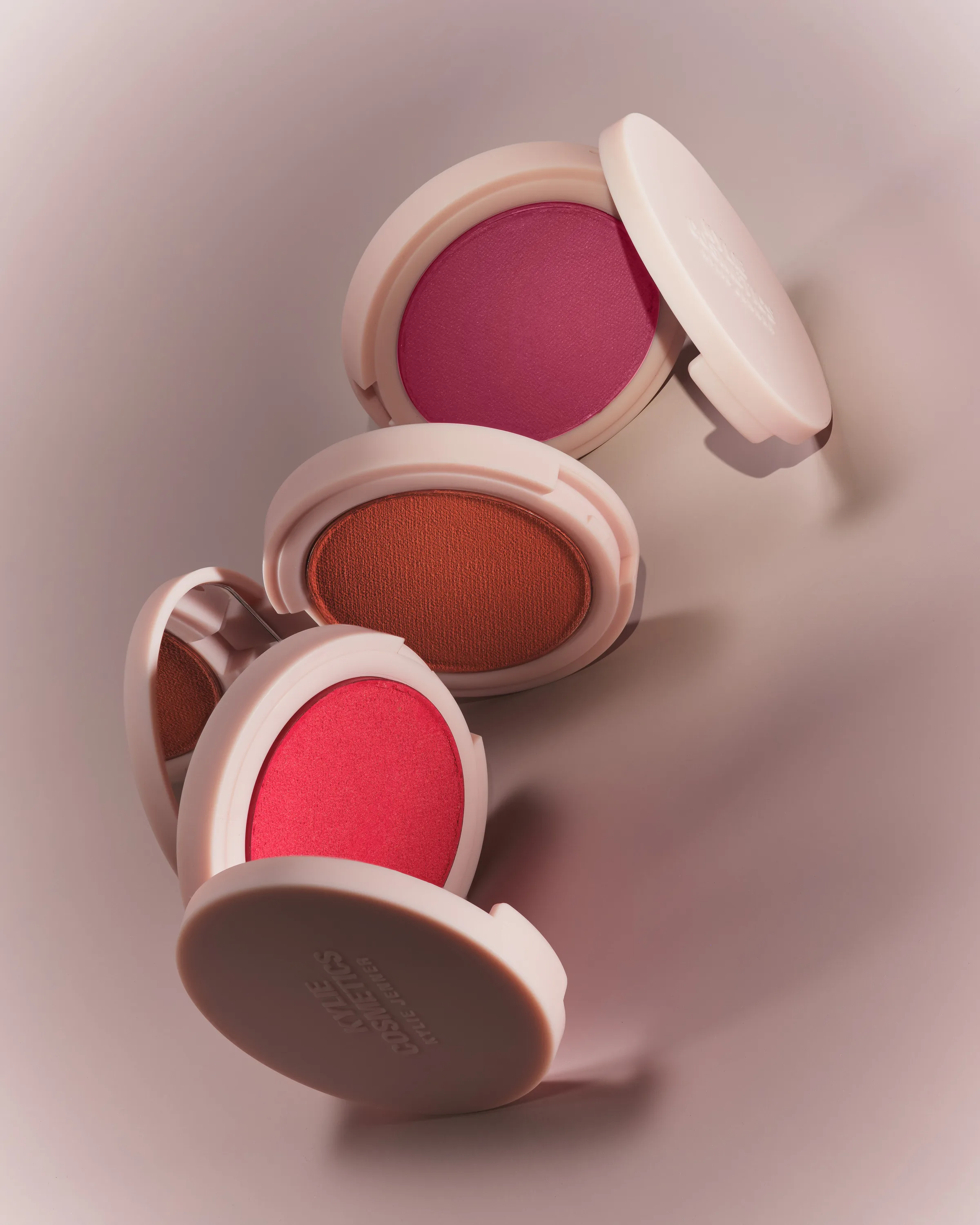 Three open pale pink blush compacts in berry, sienna and red shades stacked on a pale pink-grey backdrop, for Kylie Cosmetics.