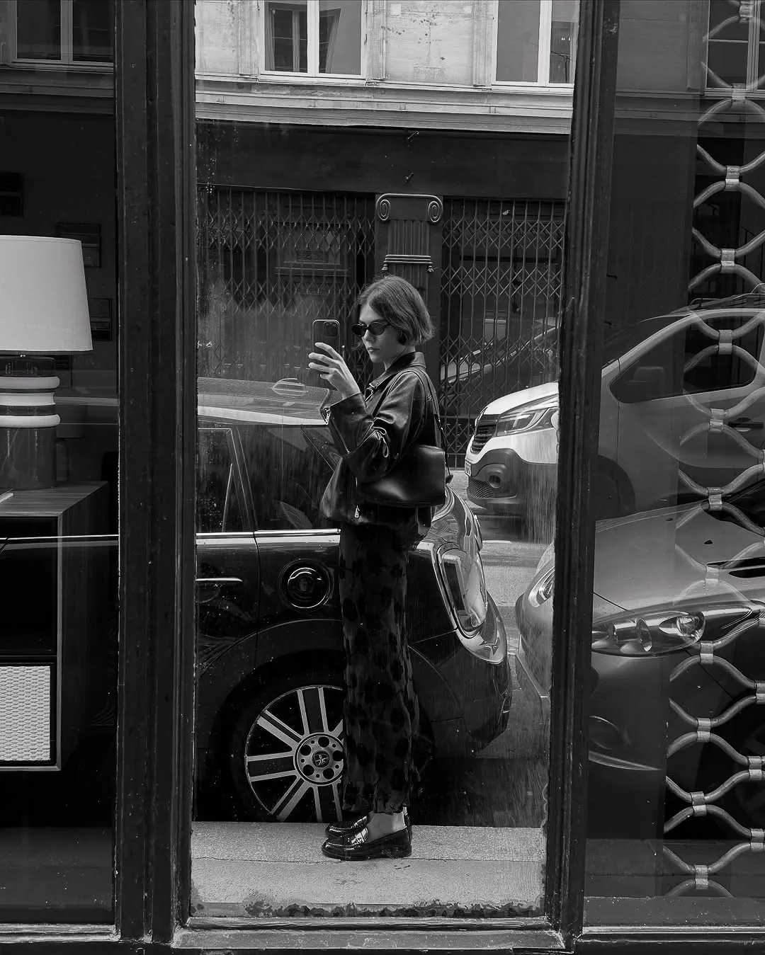 Black and white mirror selfie of a woman in sunglasses, leather jacket and loafers reflected in a street shop window.