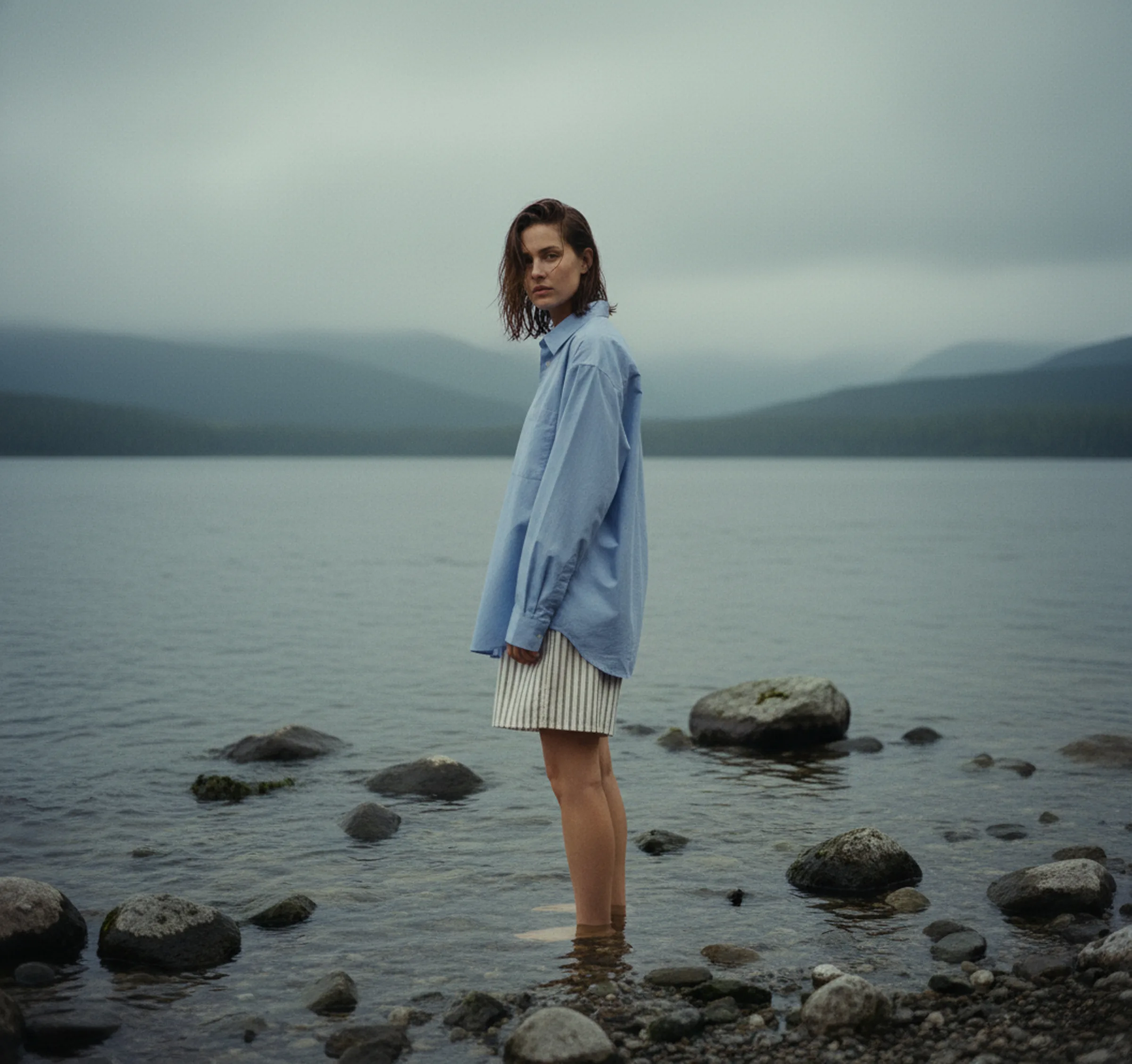 AI image of a model in an oversized blue shirt and striped shorts standing in a misty mountain lake. Tekla AI Concept.