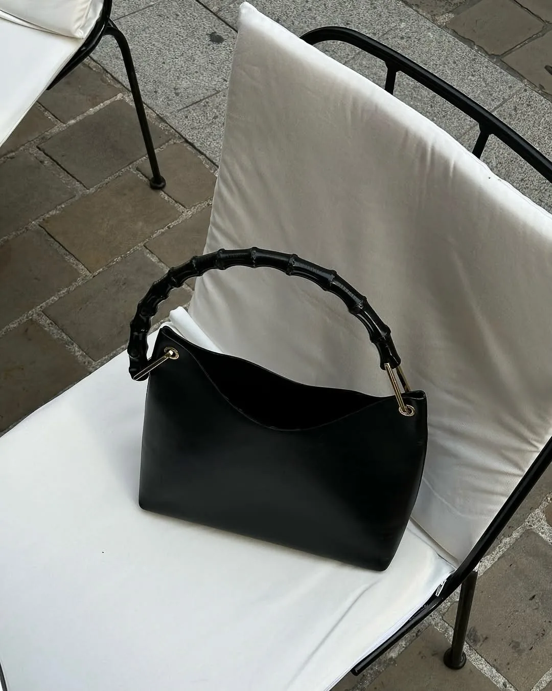 Black leather handbag with a bamboo top handle resting on a white-cushioned café chair outdoors, for Aina Velasco.