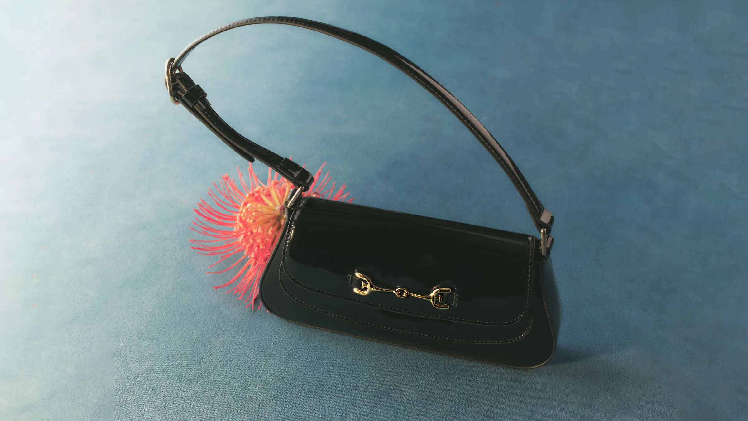 Black patent leather shoulder bag with gold horsebit hardware beside a pink pincushion protea on blue velvet, for Sam Edelman.