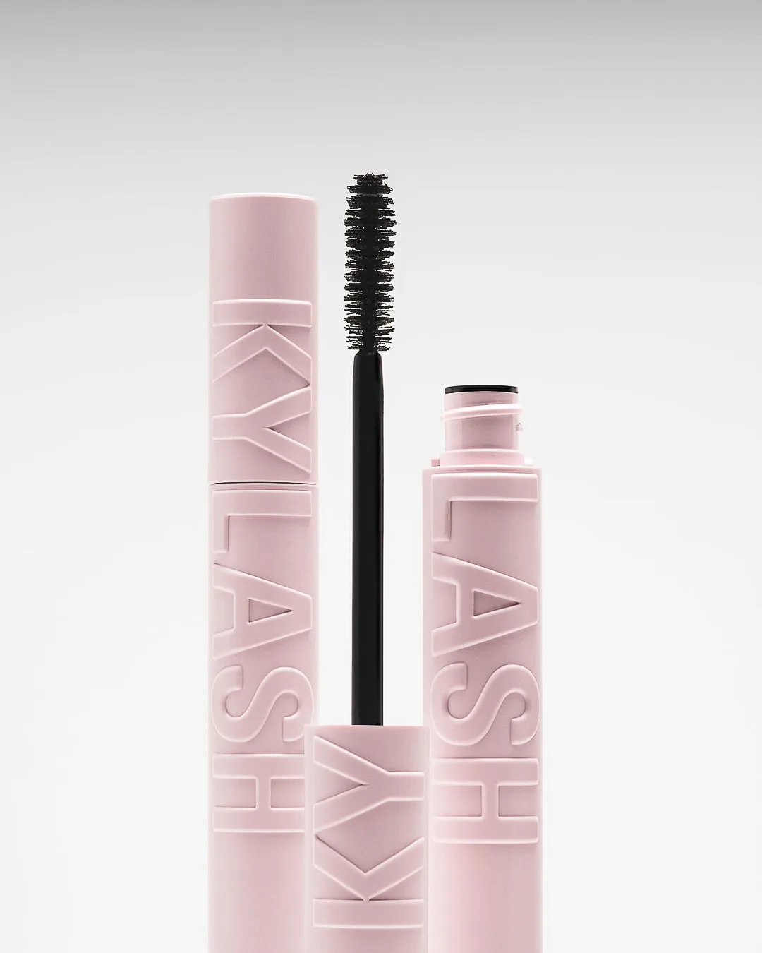 Pink Kylash mascara tubes with embossed lettering and an upright black wand on a soft grey backdrop, for Kylie Cosmetics.