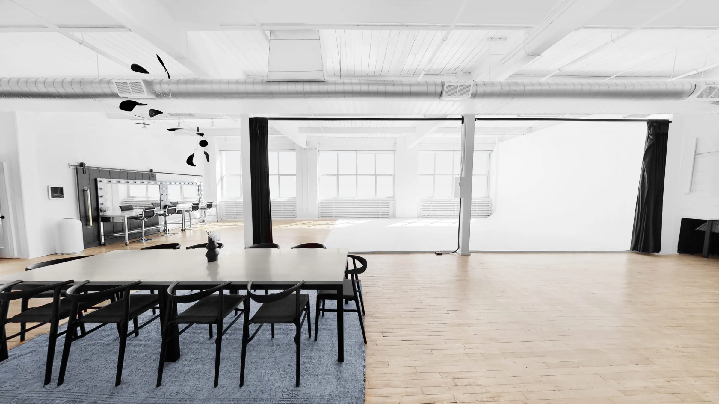 Studio 1 with a long dining table and black chairs, a makeup station and a white cyclorama under large windows.