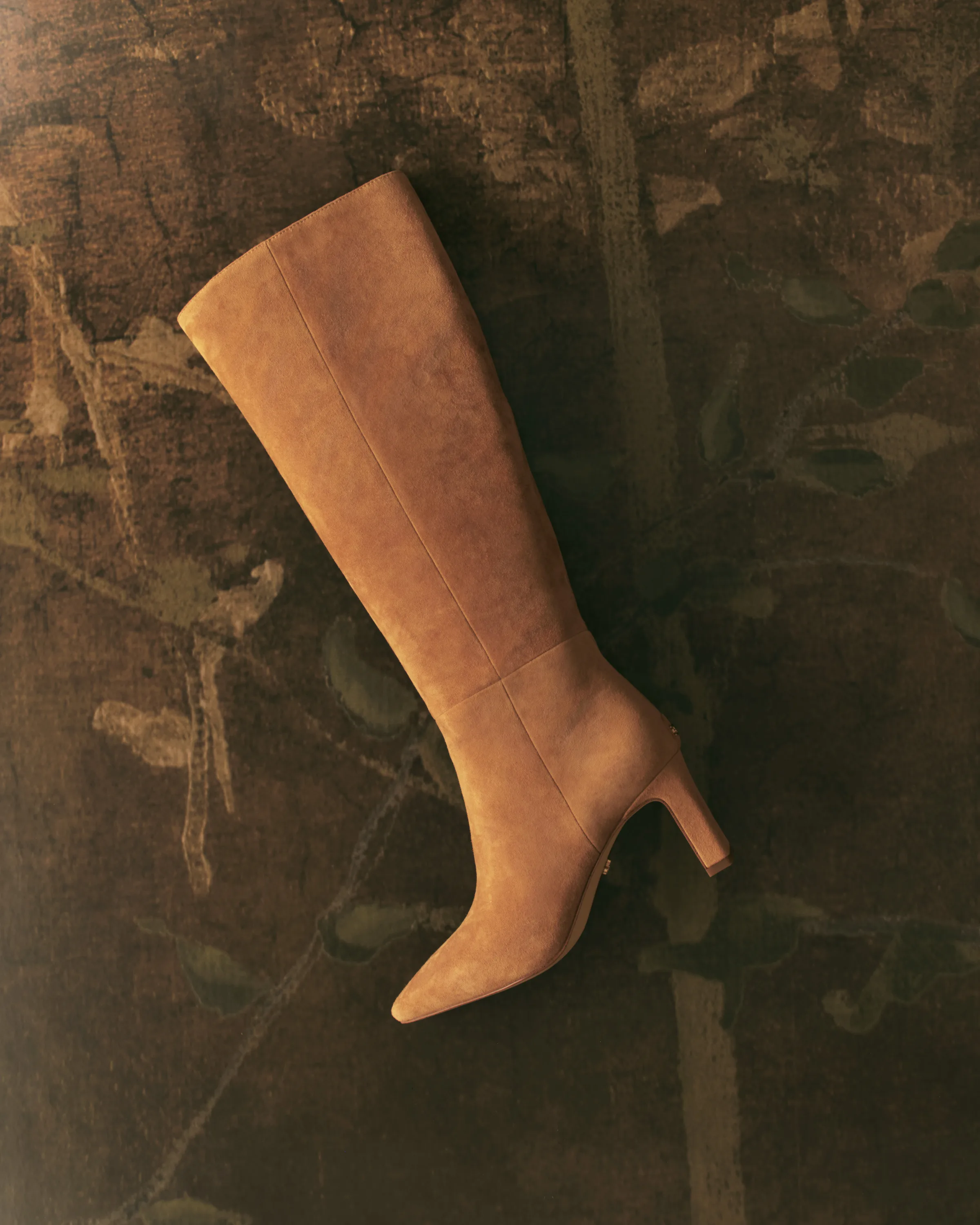 Tan suede knee-high boot with pointed toe and slim heel laid on a dark botanical tapestry, for Sam Edelman.