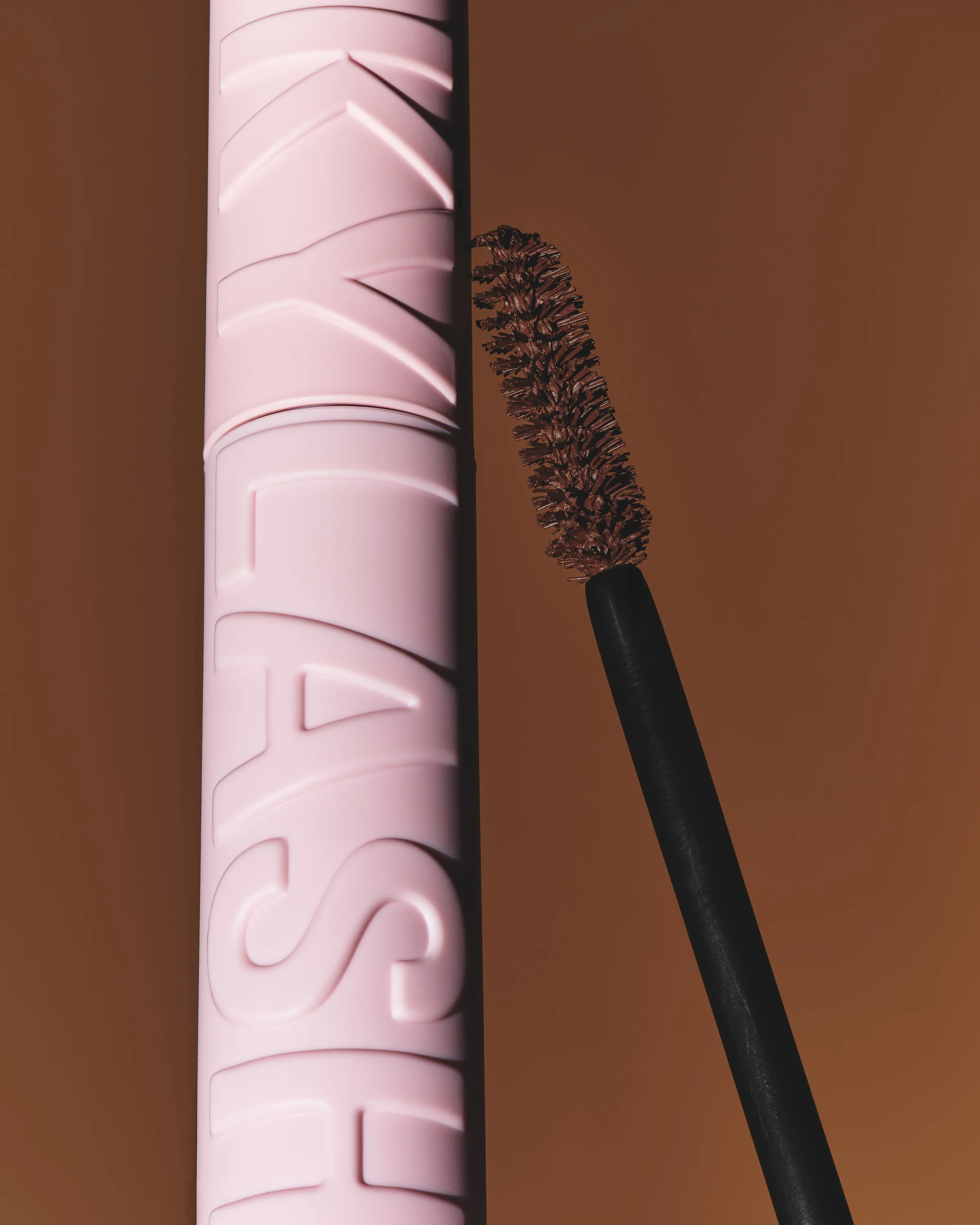 Pink embossed Kylash mascara tube beside a brown-bristled wand on a warm brown backdrop, for Kylie Cosmetics.