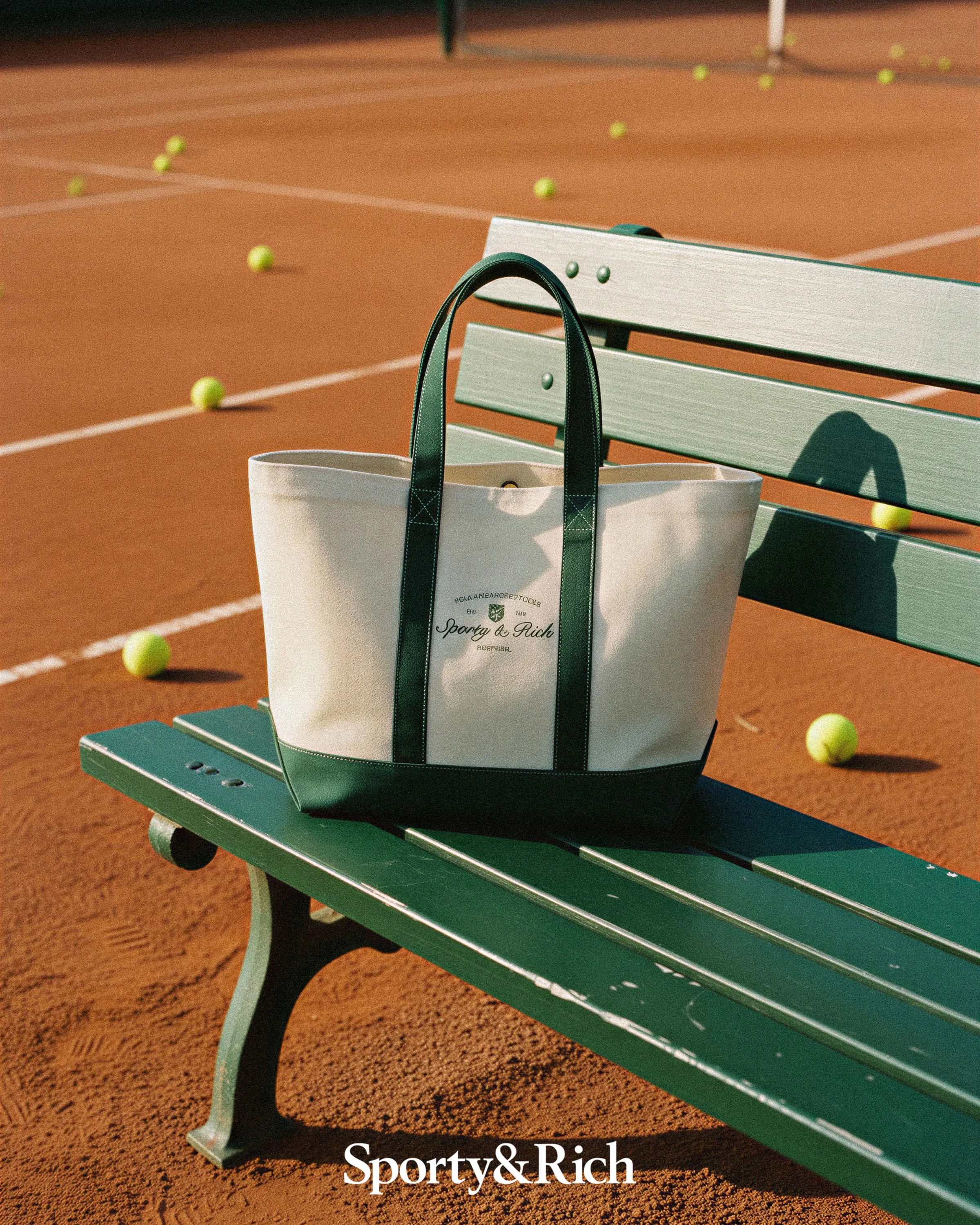 AI image of a cream canvas tote with green handles on a green bench beside a clay tennis court with balls. Sporty & Rich AI Concept.