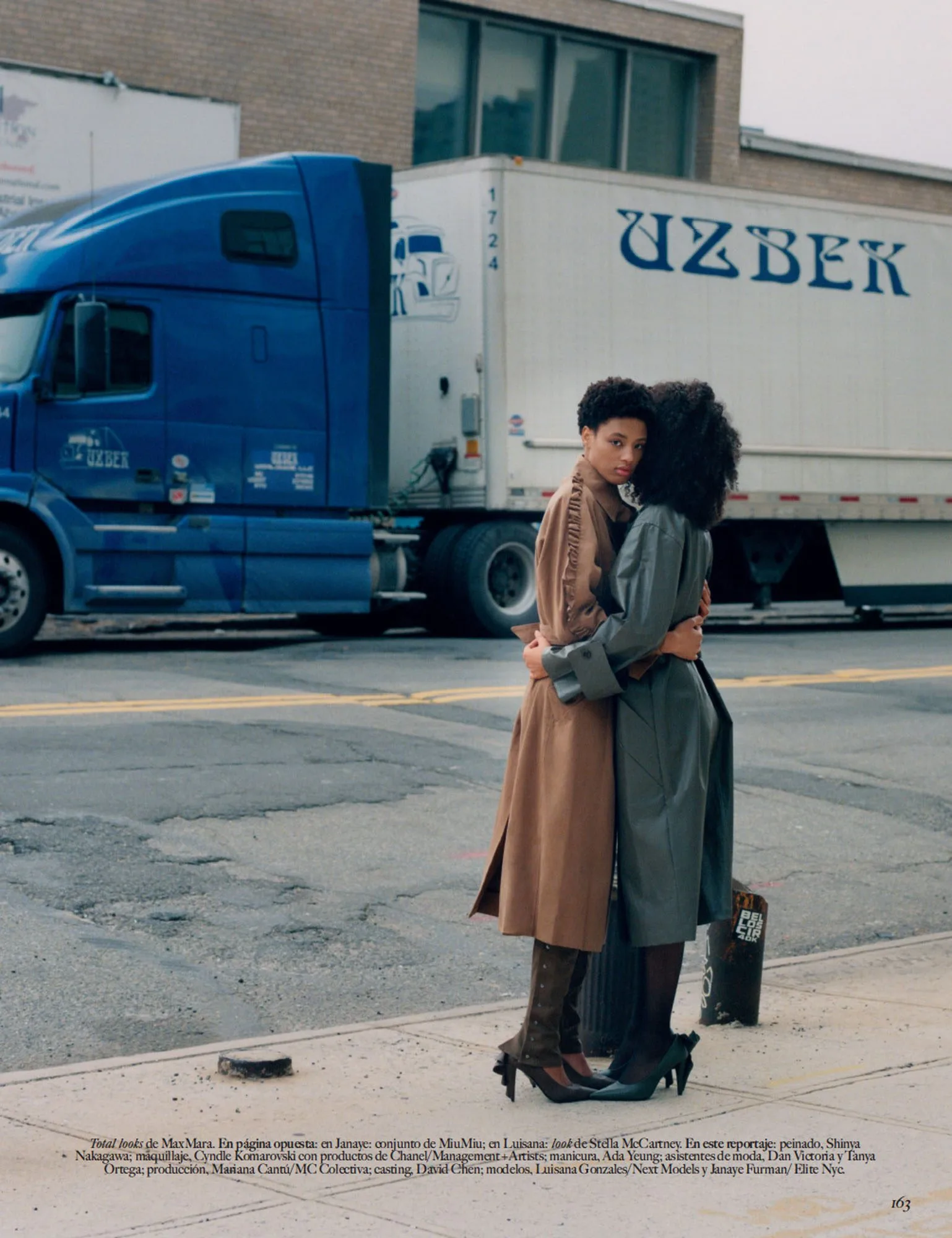 Two models in camel and grey trench coats embrace on a street before a blue truck, a magazine page, for Vogue Mexico.
