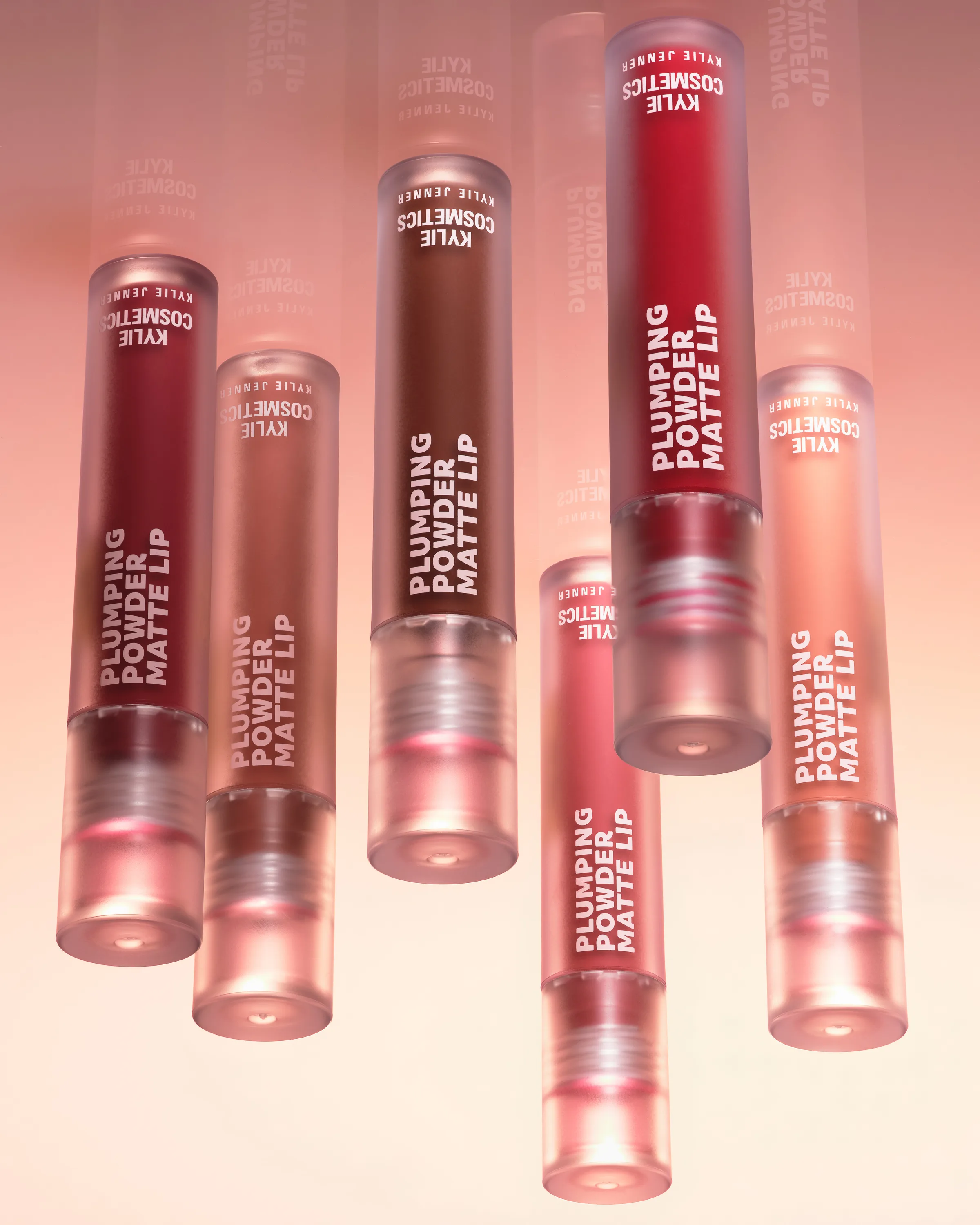 Six frosted Plumping Powder Matte Lip tubes in red, brown and nude shades suspended against a peach backdrop, for Kylie Cosmetics.