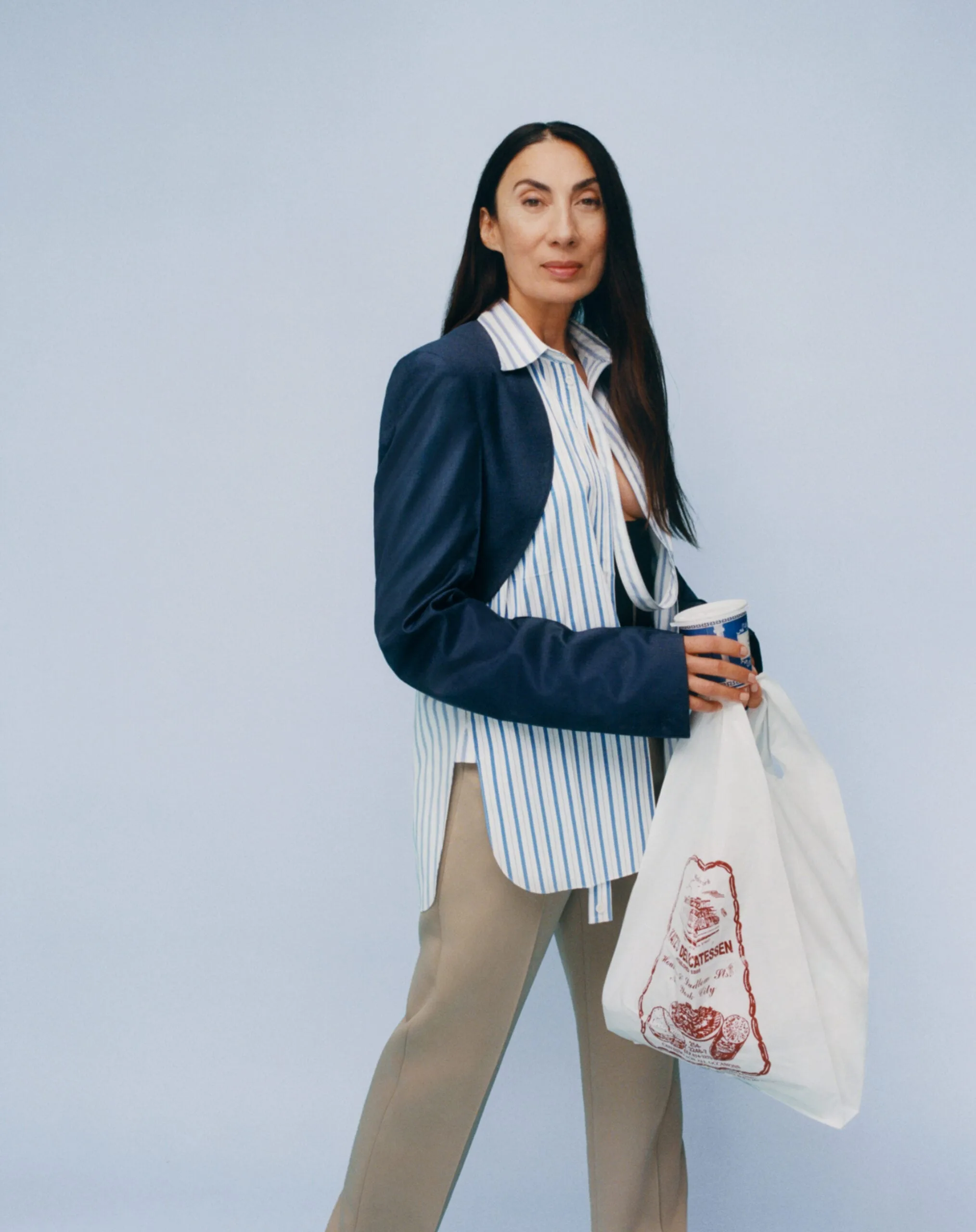 Woman in navy blazer, striped shirt and khaki trousers holding a deli bag and cup on pale blue, for Interview Magazine.