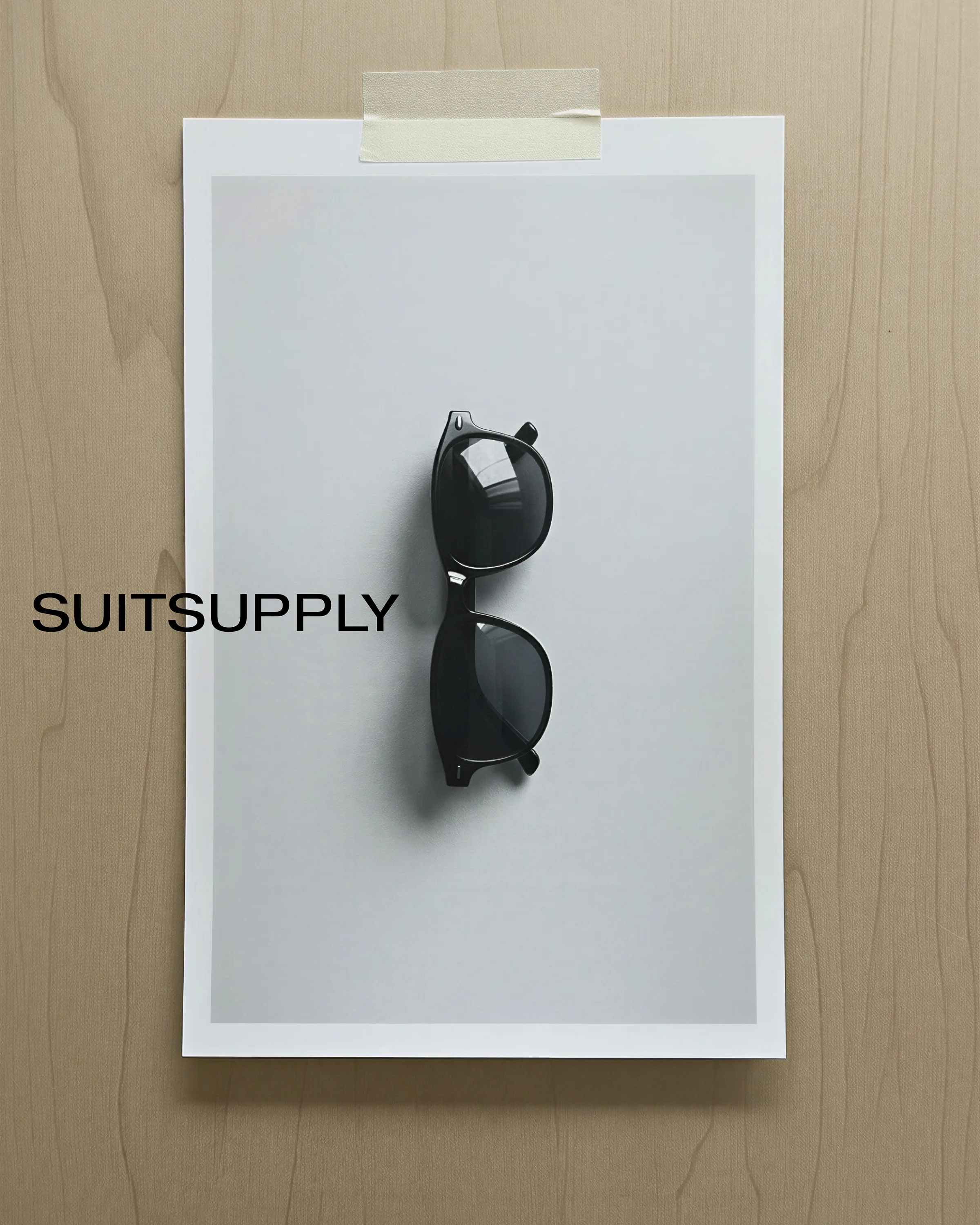 AI image of black sunglasses on a grey print taped to a wooden wall, with Suitsupply wordmark. Suitsupply AI Concept.