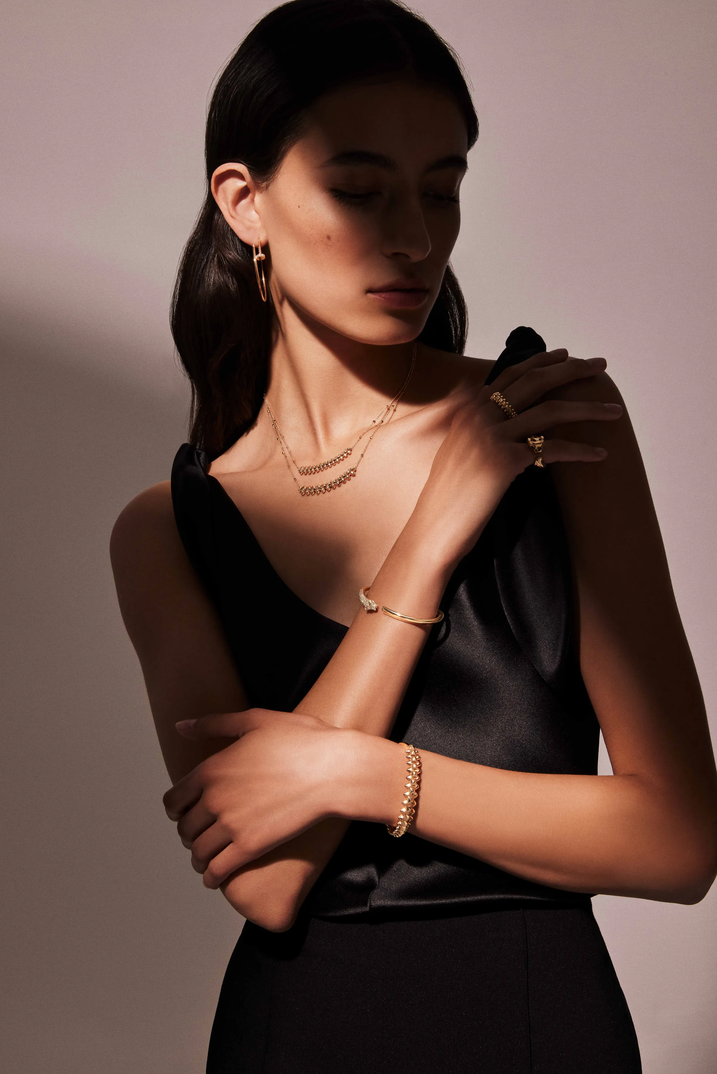 Model in a black satin dress wearing gold earrings, layered necklaces, rings and bracelets in soft shadow, for Cartier.