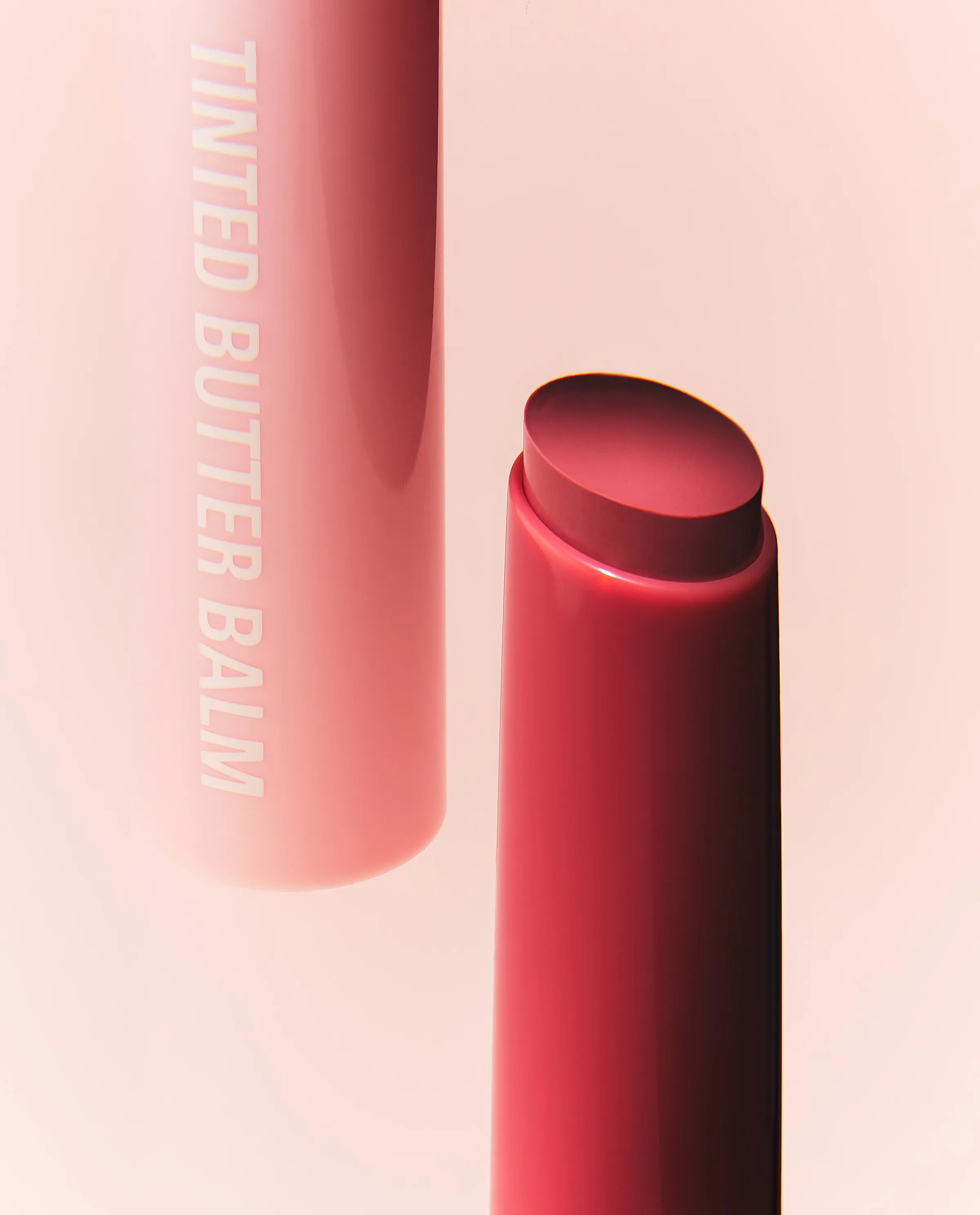 Still life of a pink Tinted Butter Balm tube and an uncapped red balm stick on a soft pink backdrop, for Kylie Cosmetics.