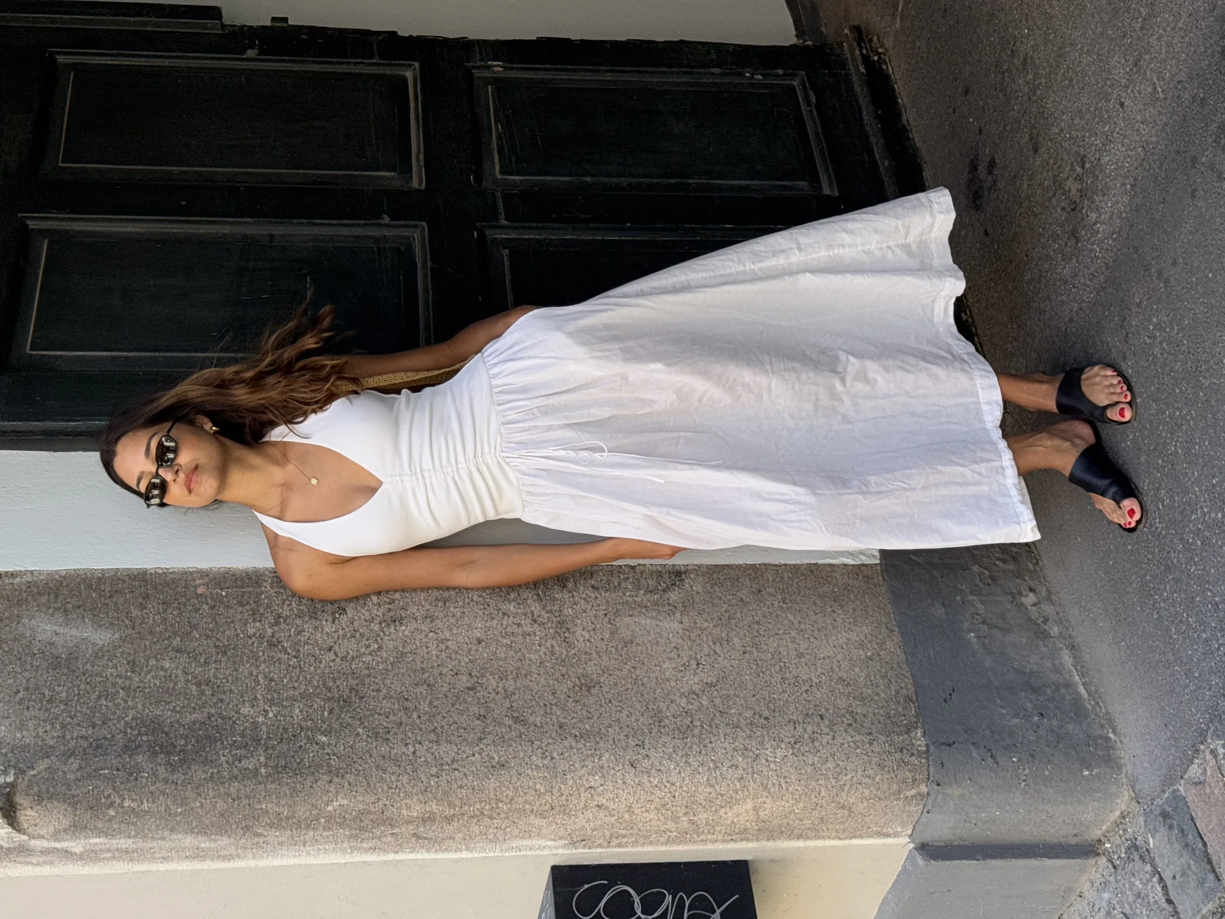 Woman in a white halter midi dress, sunglasses and black slide sandals leaning against a stone wall by a dark door.