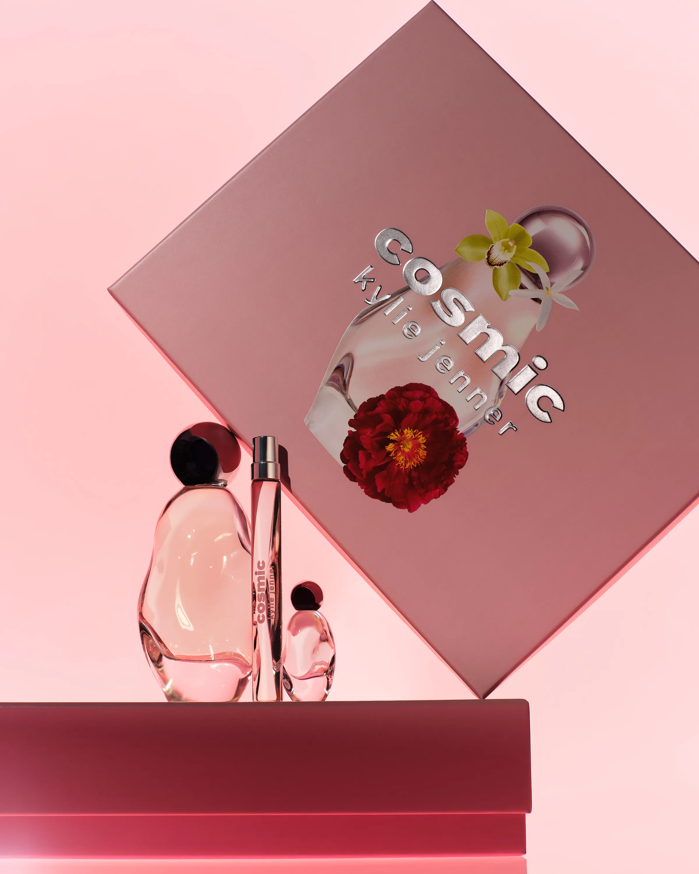 Cosmic perfume bottles, travel spray and mini on pink plinths before a tilted gift box, for Kylie Cosmetics.