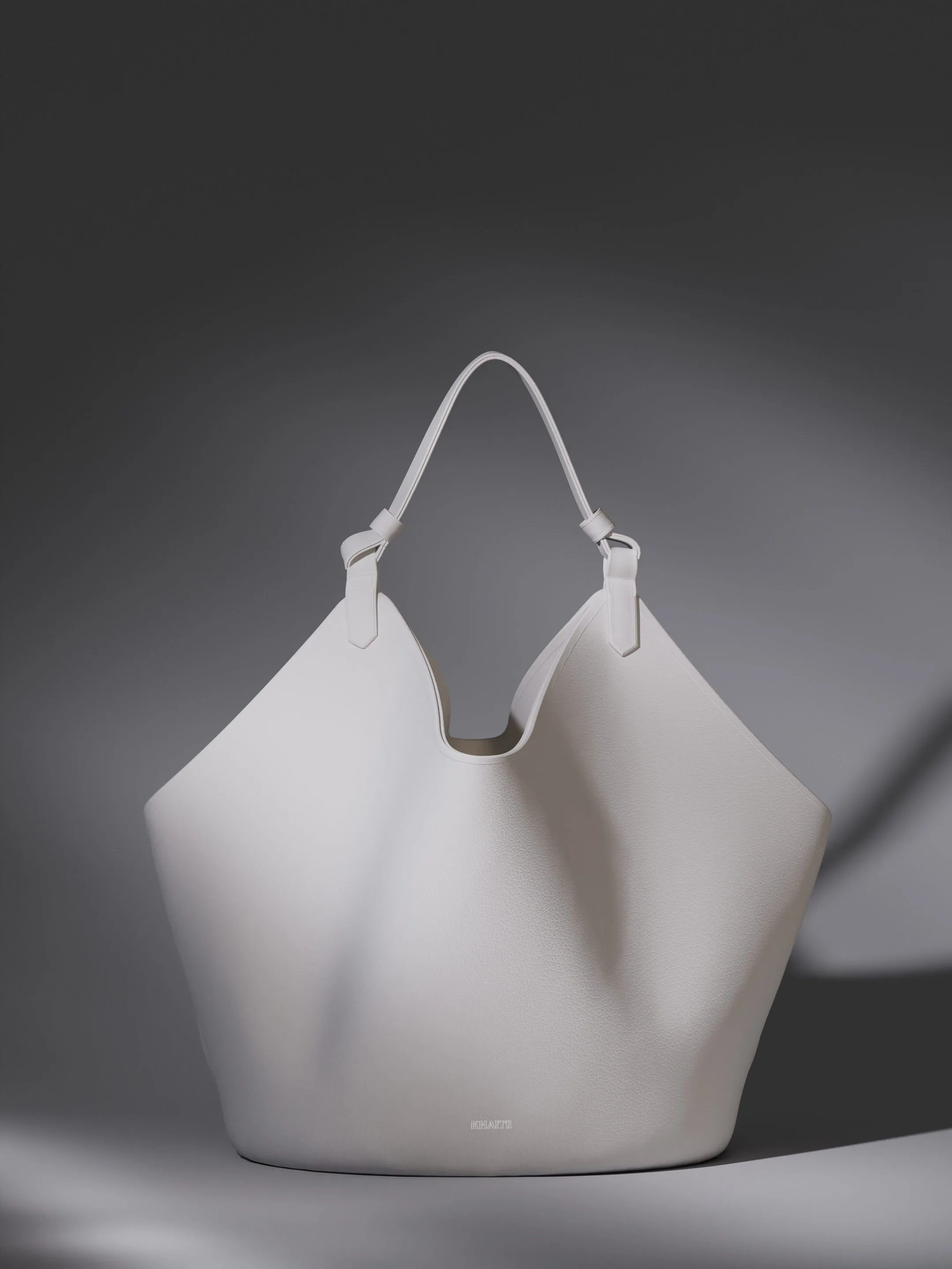 CGI render of a white grained leather tote bag with knotted strap against a grey shadowed backdrop, for Khaite.