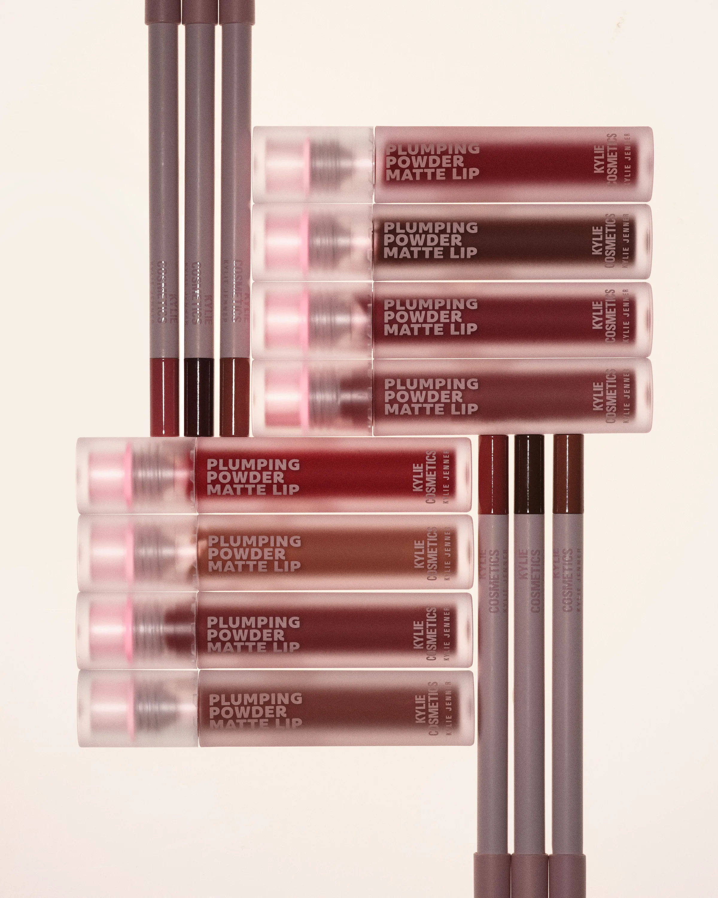 Plumping Powder Matte Lip tubes and lip liners in red and brown shades stacked on a cream background, for Kylie Cosmetics.