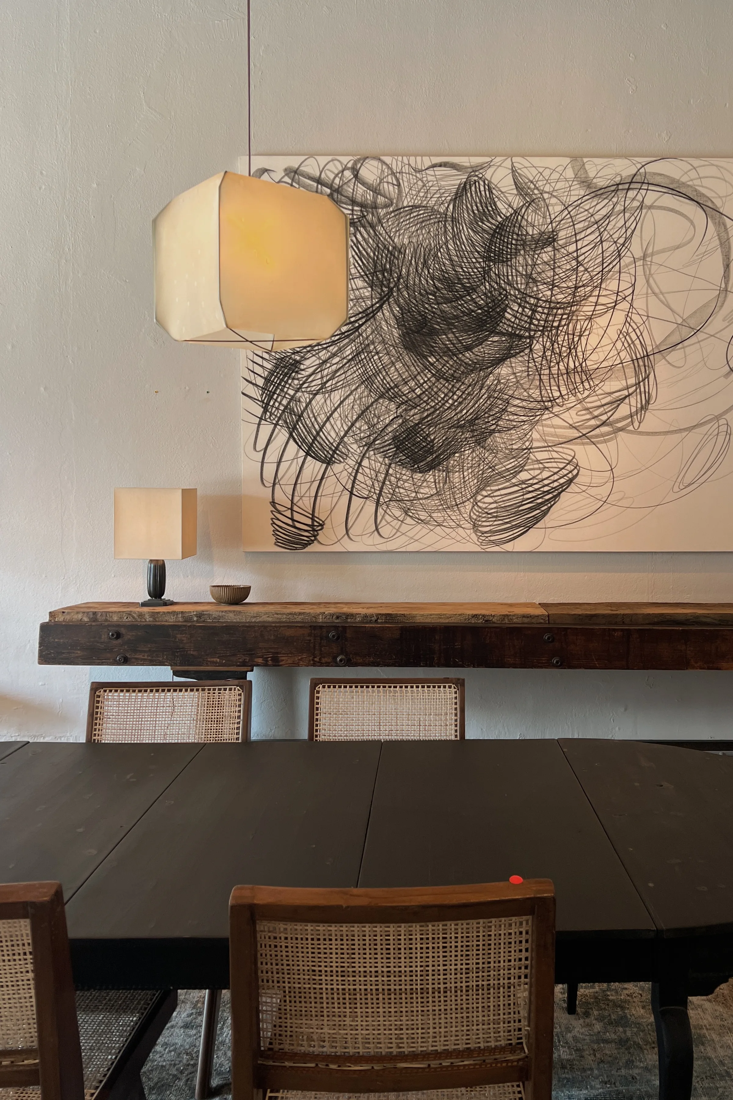 Dark dining table with cane chairs, paper pendant lamp and abstract charcoal artwork above a rustic console, for Jennifer Hagler.