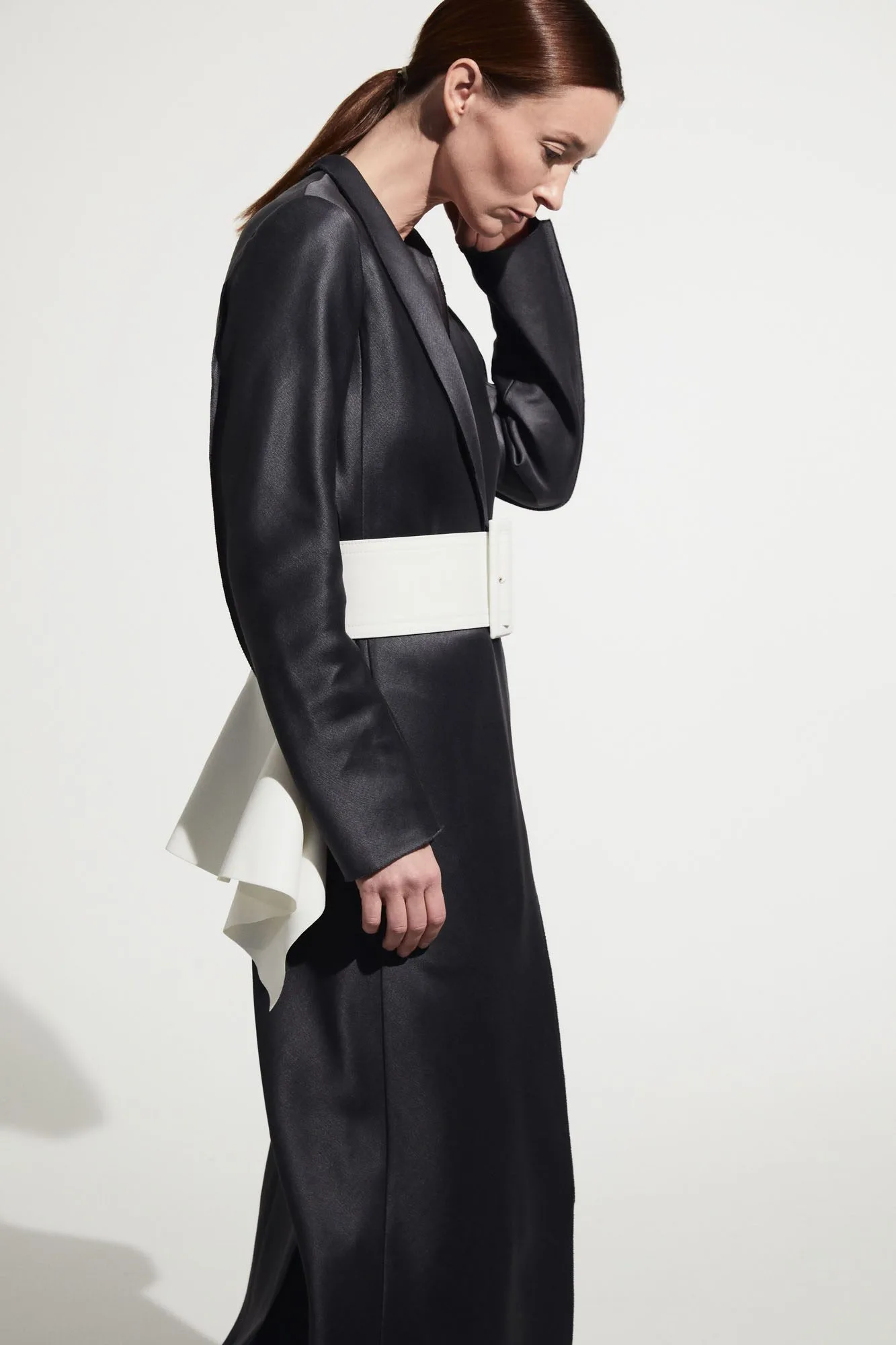 Model in a long black satin coat cinched with a wide white belt and ruffle, side profile on white, for The RealReal.
