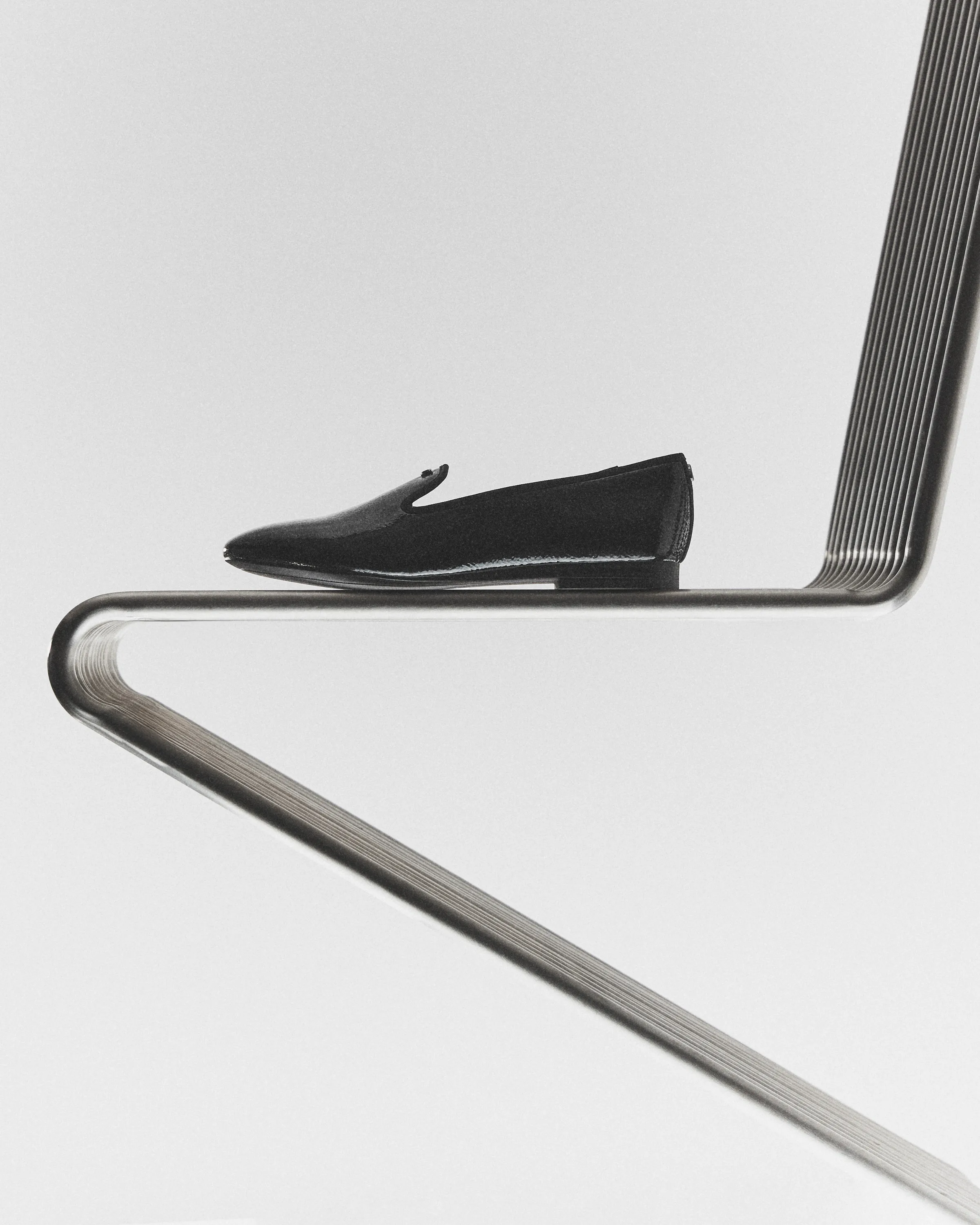 Black patent leather loafer resting on a cantilevered chrome tube chair against a pale grey backdrop, for Sam Edelman.