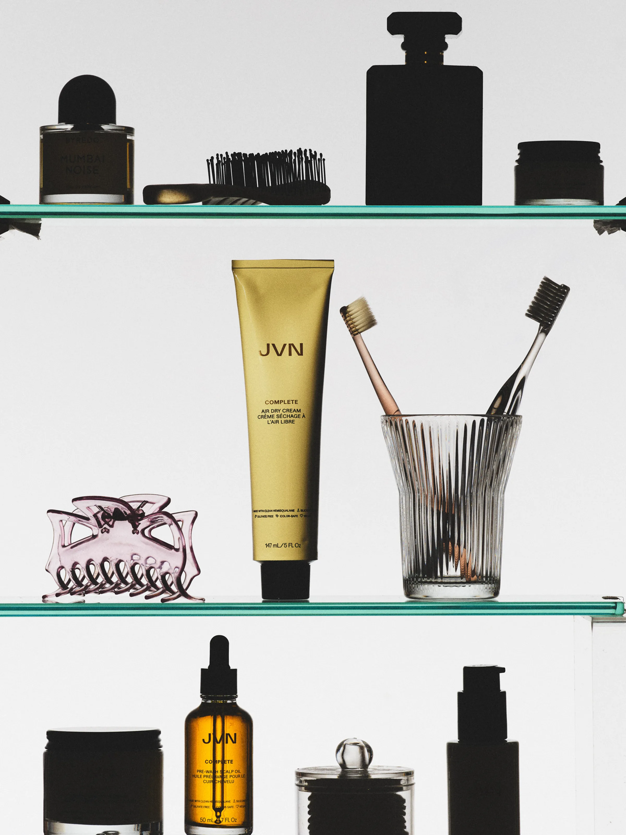 Gold air dry cream tube and hair oil on backlit glass shelves with toothbrushes, claw clip and silhouetted bottles, for JVN.