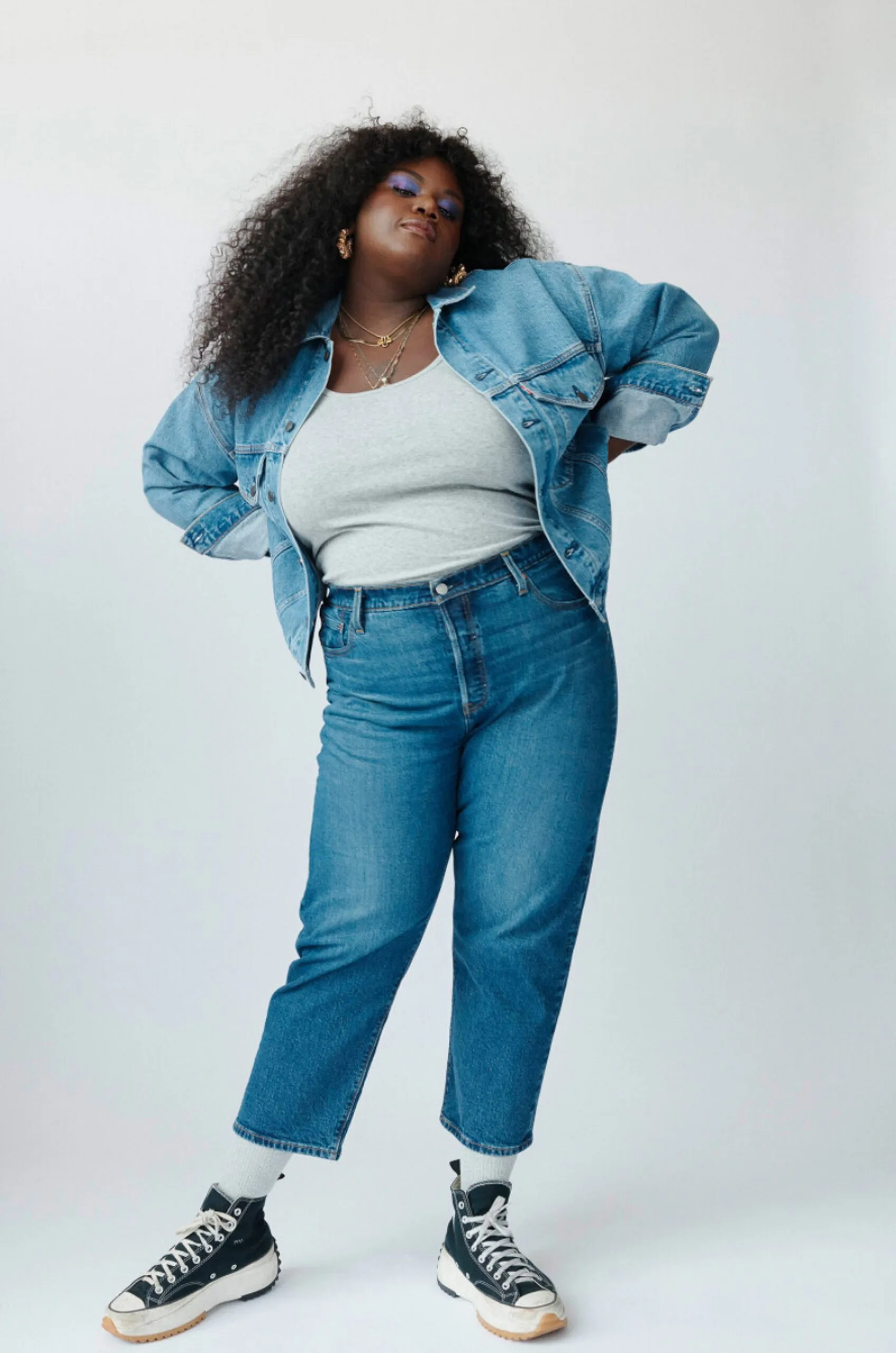 Model in a denim trucker jacket, grey tank and cropped blue jeans with black platform sneakers on a white backdrop, for Levi's.