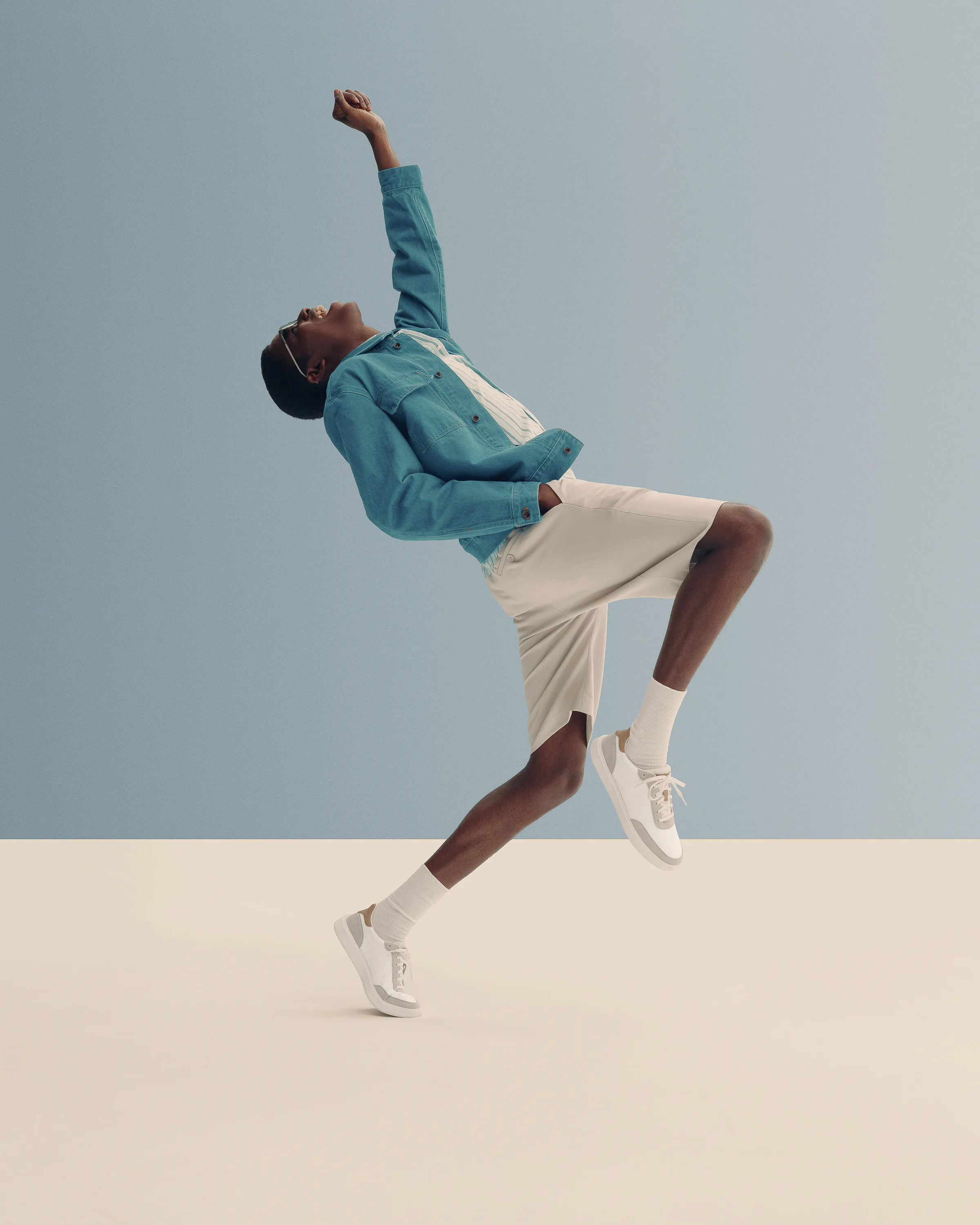 Model leaping with fist raised in teal denim jacket, cream shorts and white sneakers against a blue and beige set, for Cole Haan.