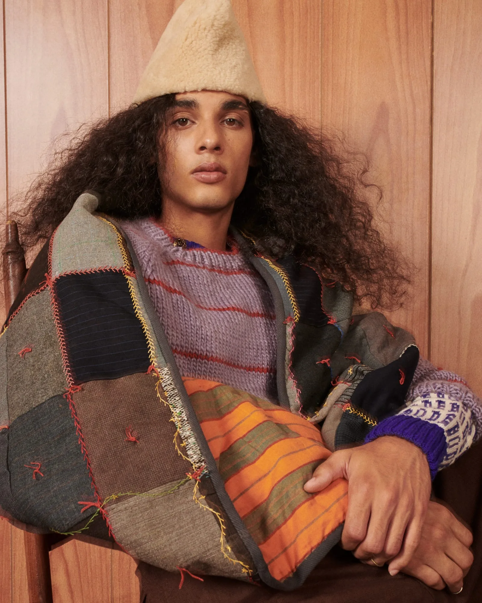 Model with long curly hair in a patchwork quilted jacket, striped knit sweater and shearling hat against wood panelling.