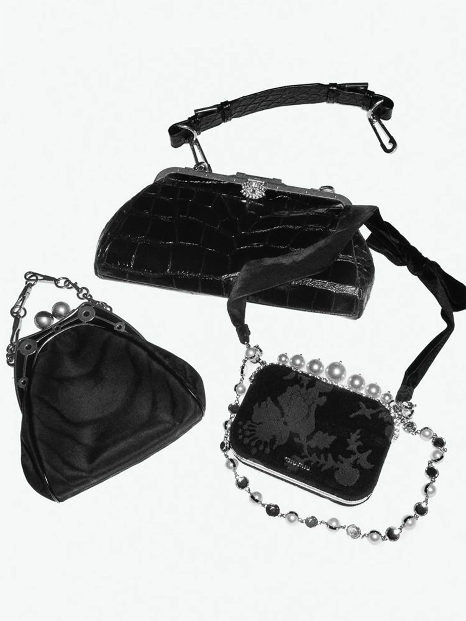 Black and white still life of three black evening bags: croc-embossed frame bag, satin pouch and pearl-chain clutch.
