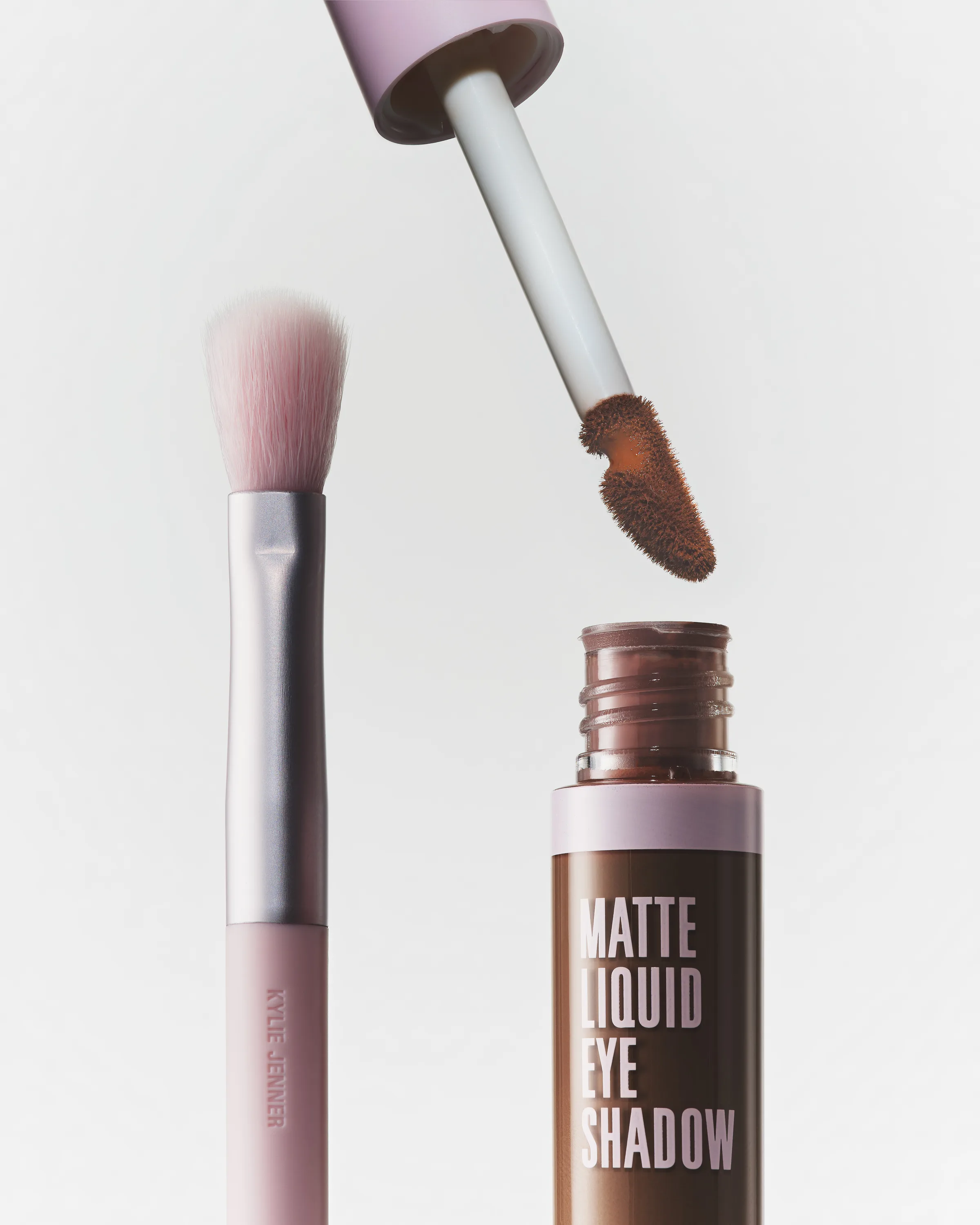 Pink eyeshadow brush beside an open brown matte liquid eyeshadow with its applicator lifted, on white, for Kylie Cosmetics.