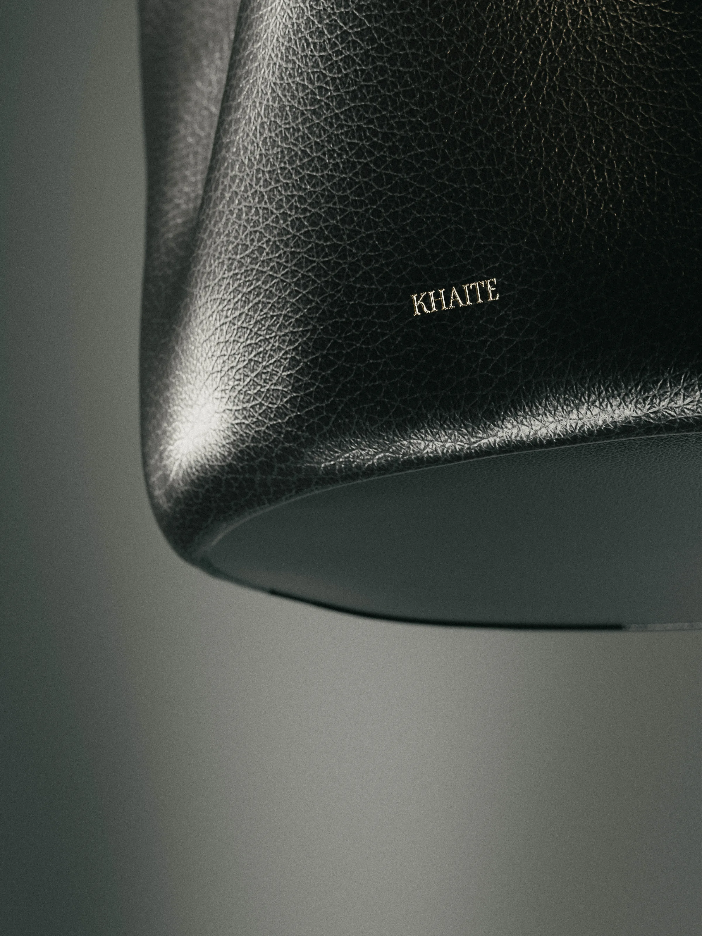 CGI render of a black pebbled leather bag base with a gold-stamped logo against a grey backdrop, for Khaite.
