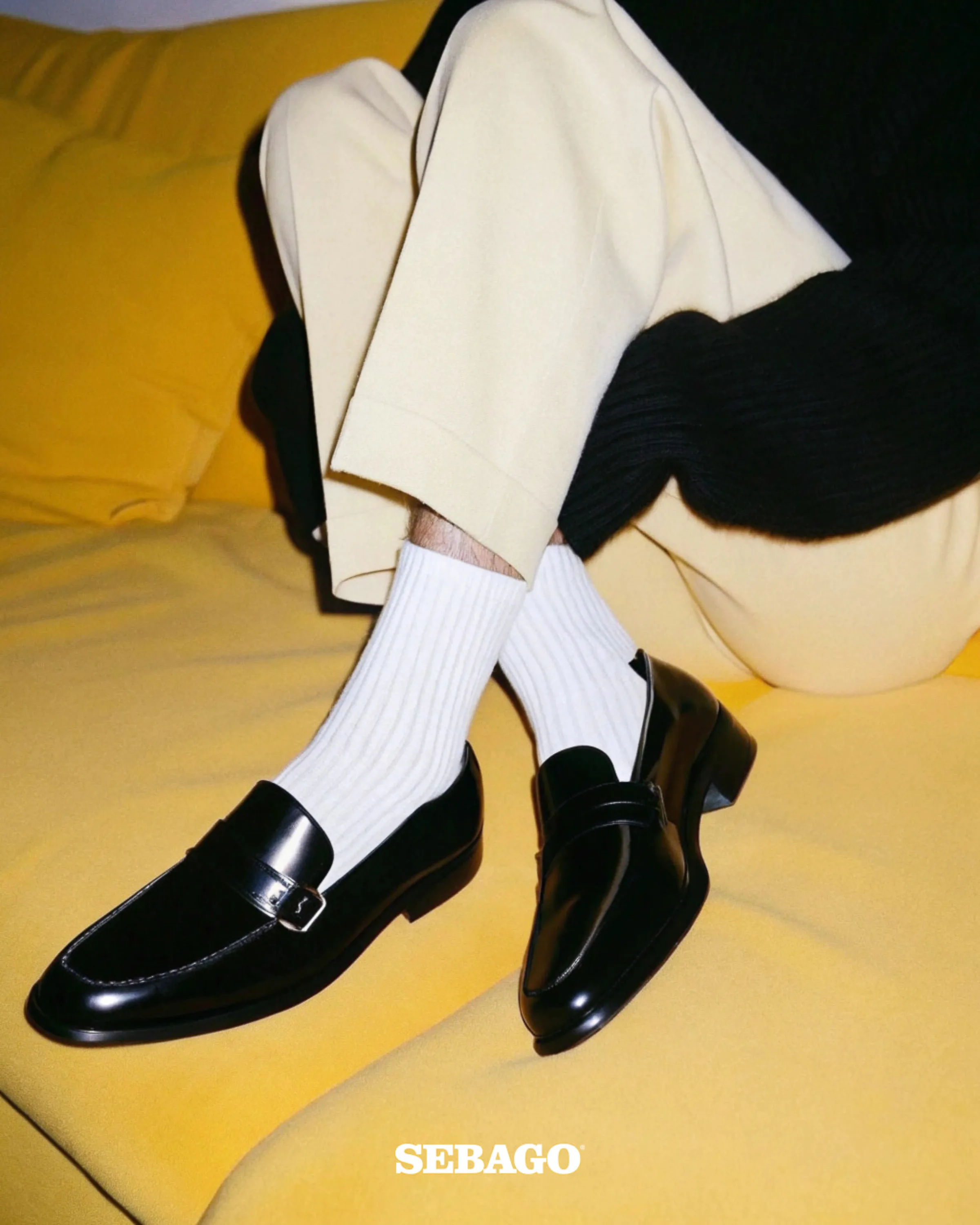 AI image of black leather buckle loafers with white ribbed socks and cream trousers on a yellow sofa. Sebago AI Concept.