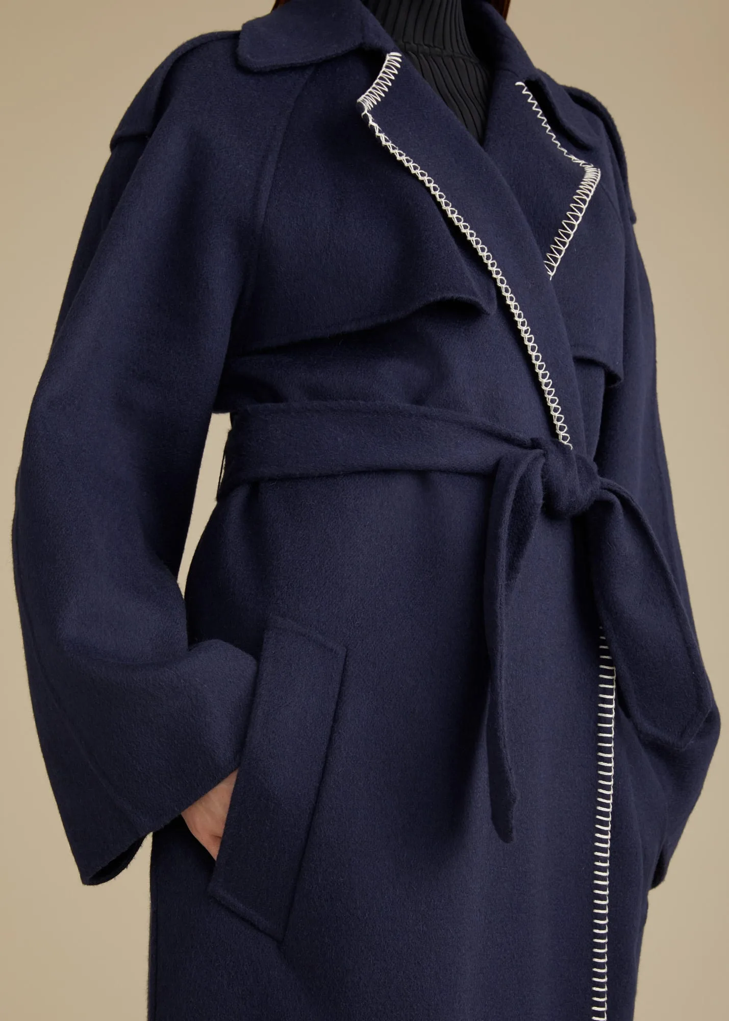 Close-up of a model in a navy belted wool coat with white whipstitch trim on the lapels, on a beige backdrop, for Khaite.