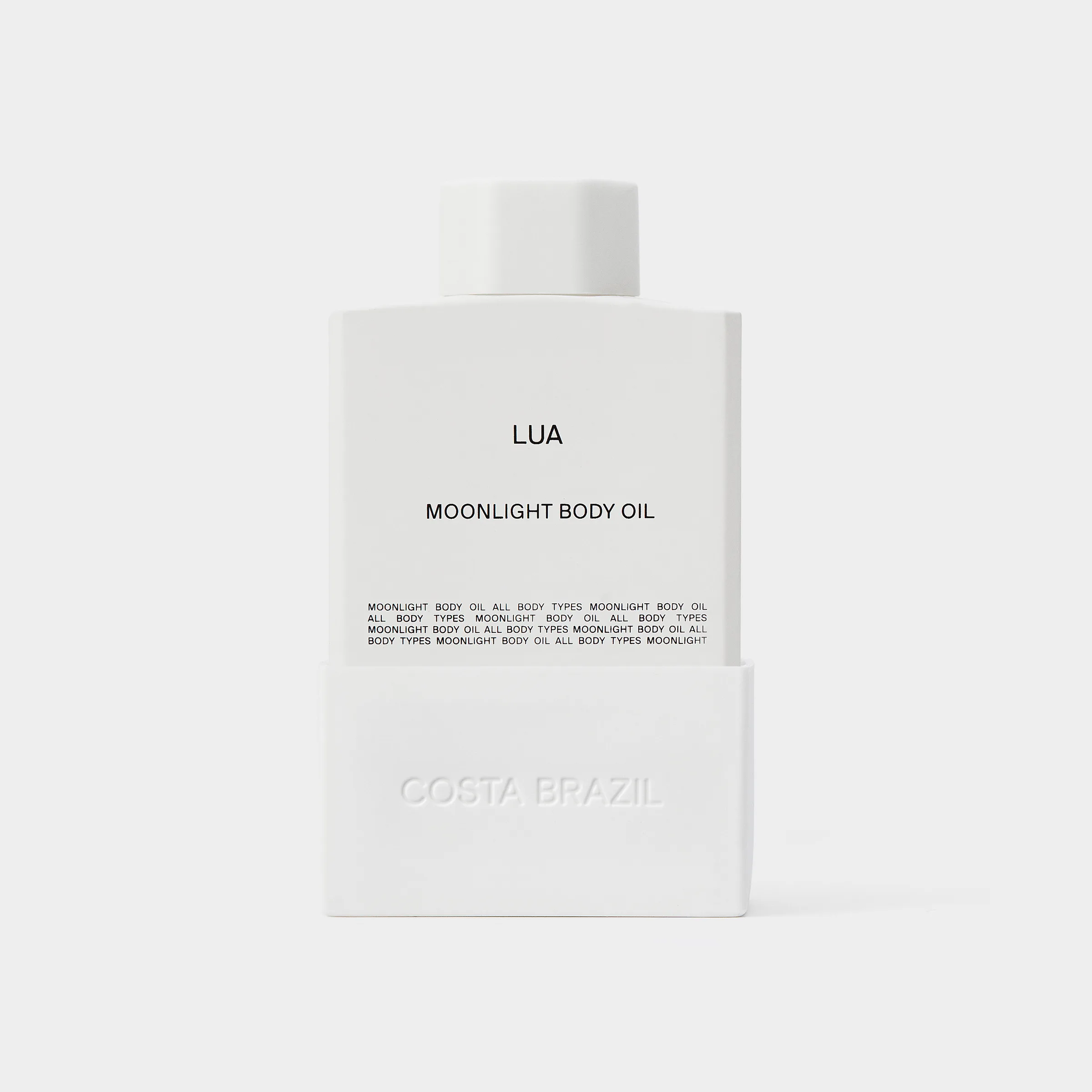 White bottle of Lua Moonlight Body Oil with embossed base on a pale grey background, for Costa Brazil.