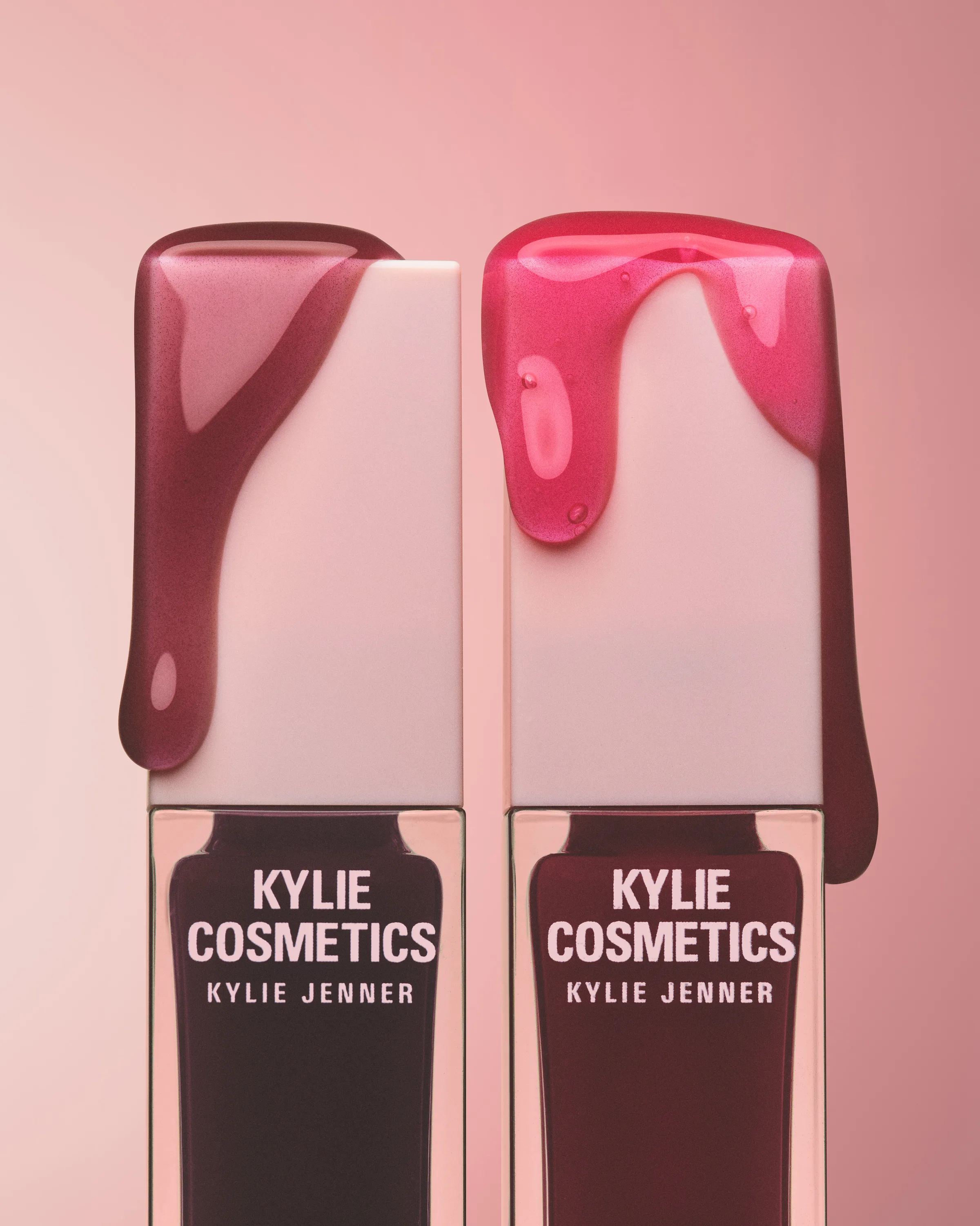 Two lip gloss tubes with berry and pink gloss dripping over their white caps on a pink background, for Kylie Cosmetics.